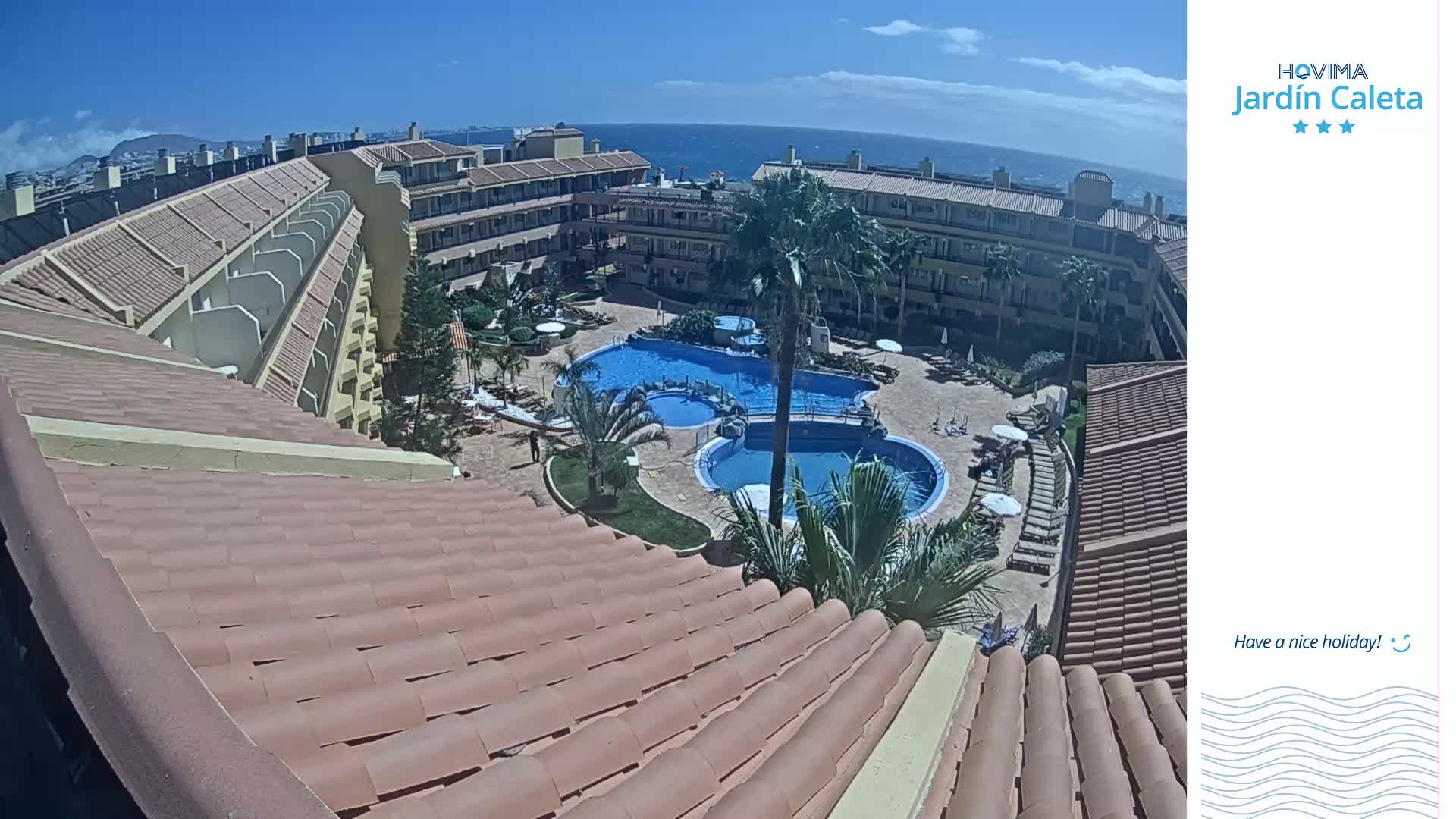 An aerial view shows a resort complex with multiple buildings surrounding two swimming pools on a sunny day, overlooking the ocean in the distance.