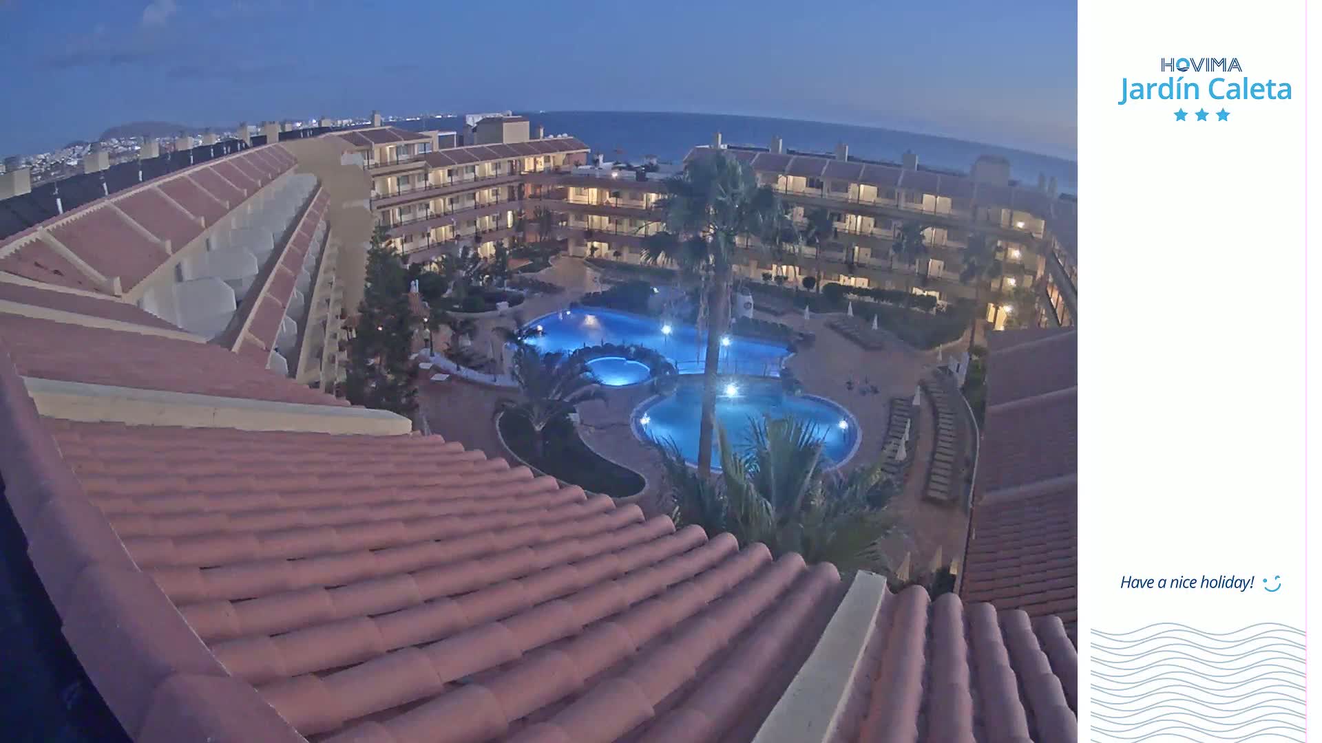 An illuminated pool complex is nestled within a low-rise building complex at dusk, overlooking a calm ocean.