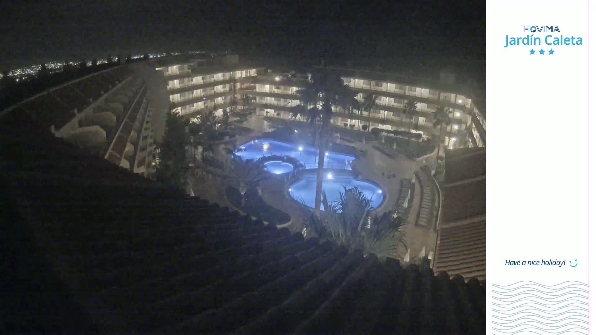 A nighttime aerial view shows a multi-story building complex surrounding illuminated swimming pools.