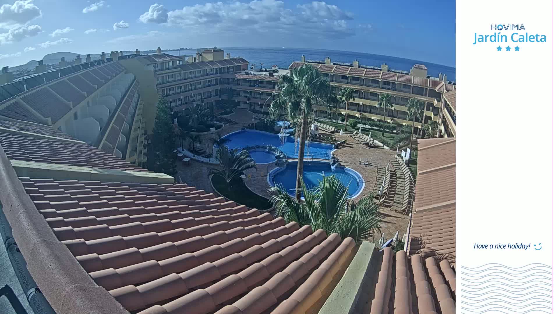 An aerial view of a resort with multiple pools, surrounding buildings, and palm trees, on a sunny day with a clear blue ocean visible in the distance.
