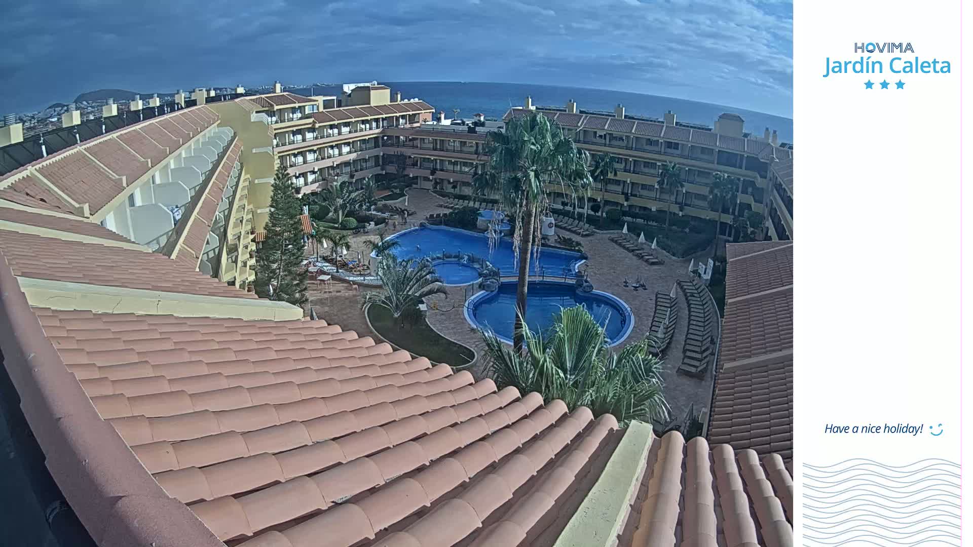 A mostly sunny day overlooks a resort with a large pool, surrounded by tan buildings and palm trees.