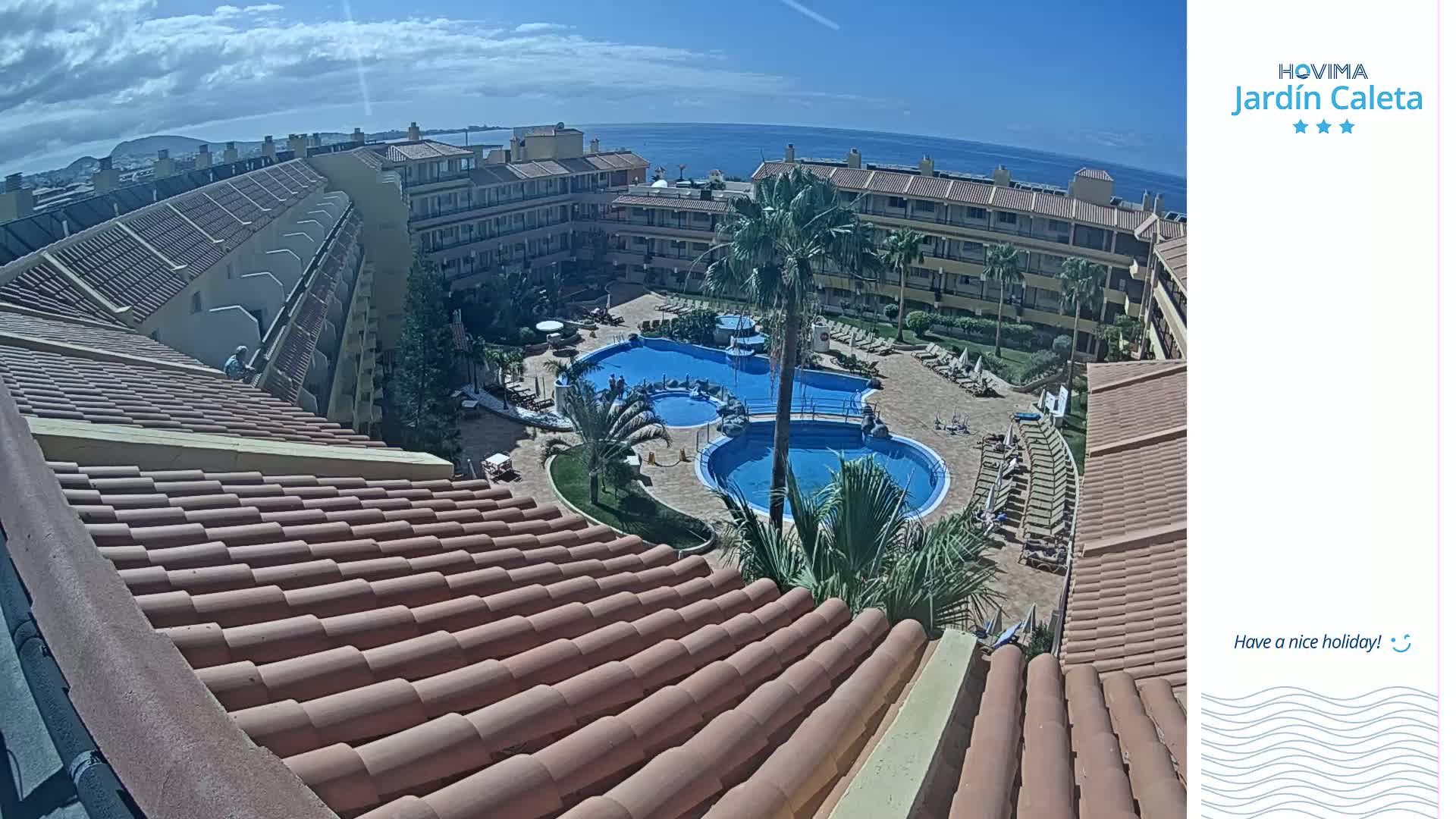 A sunny view overlooking a resort complex with multiple pools, surrounded by buildings with terracotta roofs, and an ocean in the background.