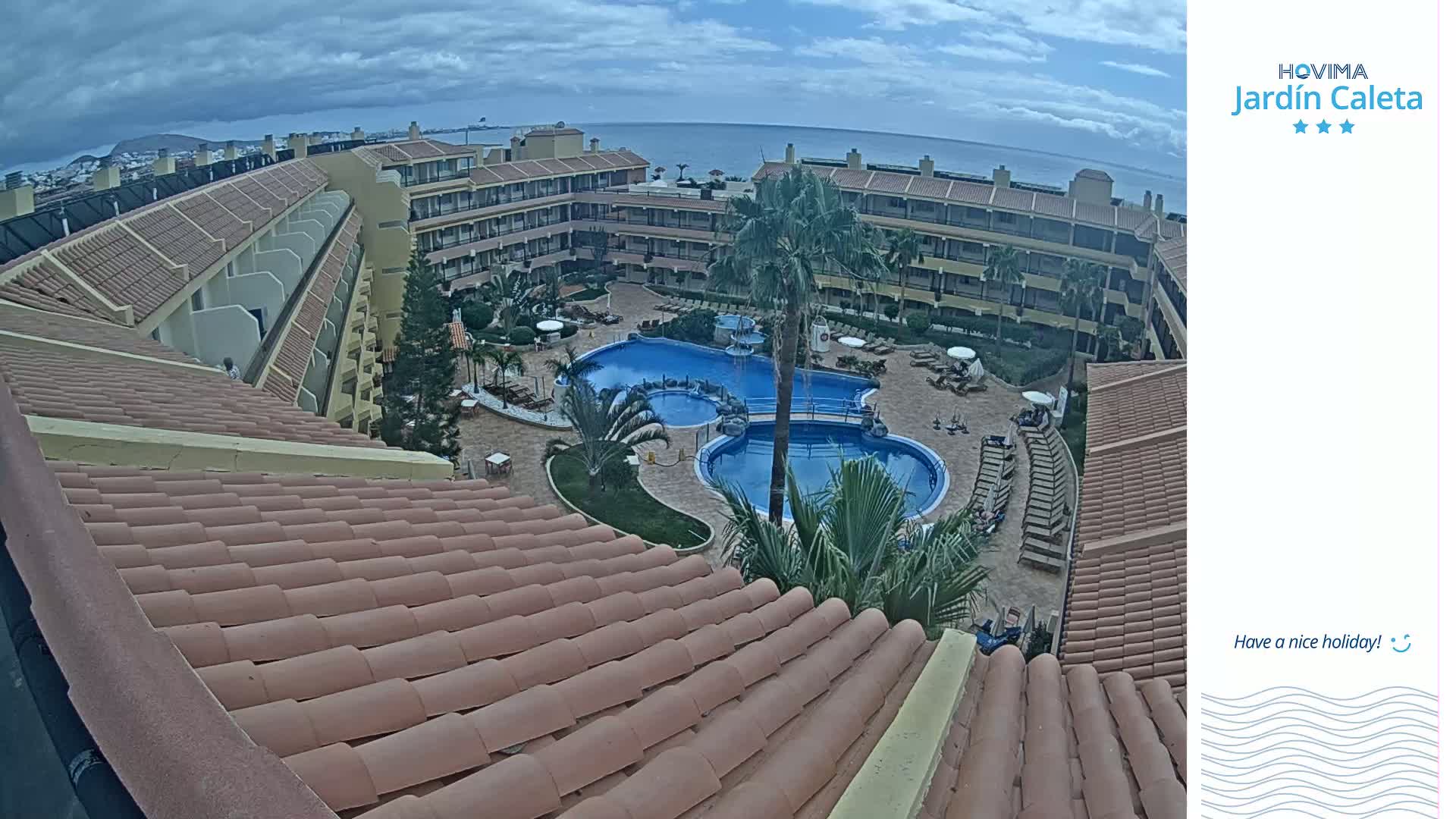 An overhead view of a resort complex with a swimming pool, under a partly cloudy sky, overlooks the ocean in the distance.