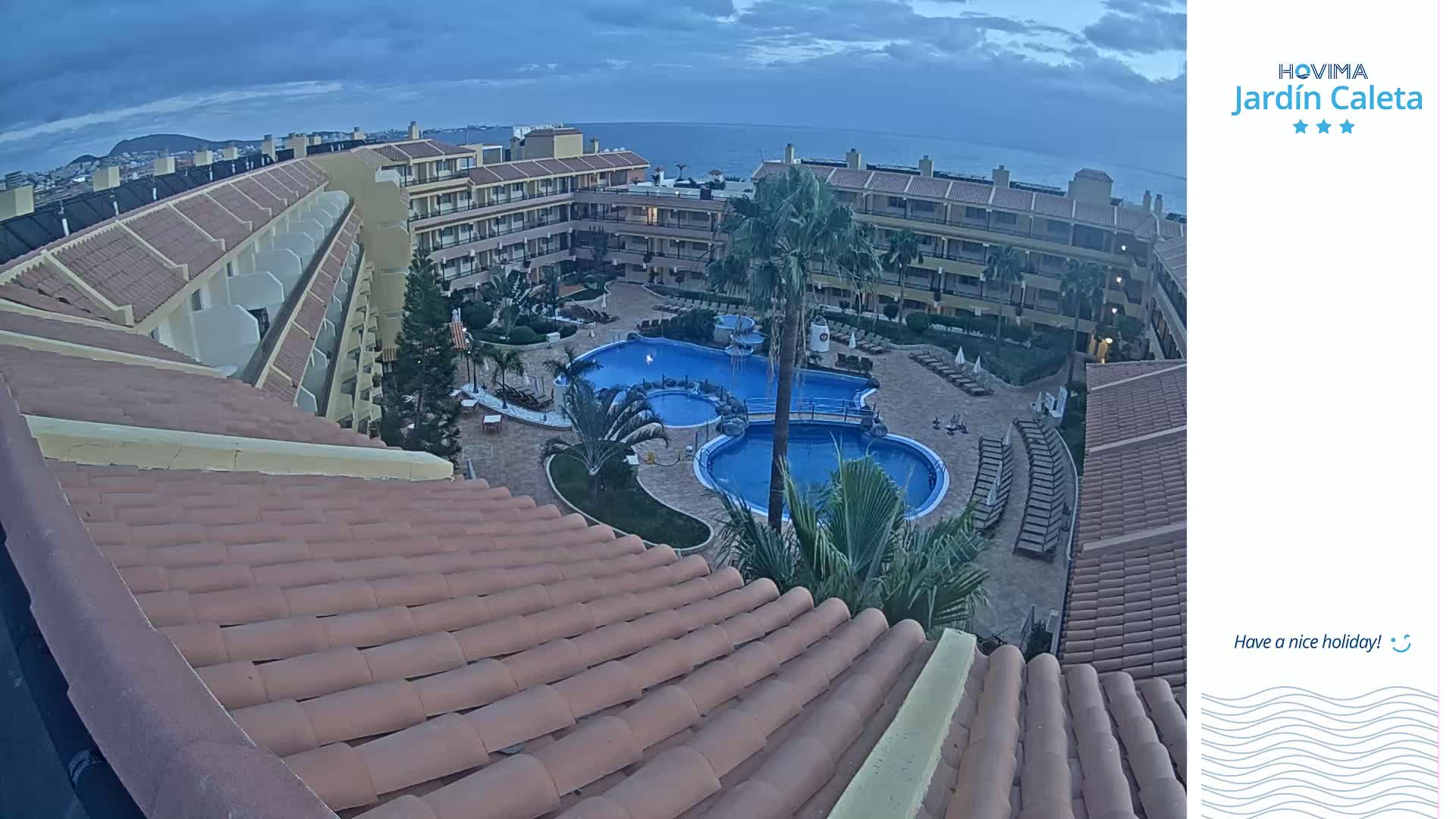 An aerial view of a resort at dusk shows a courtyard with a swimming pool, surrounded by buildings with terracotta roofs.