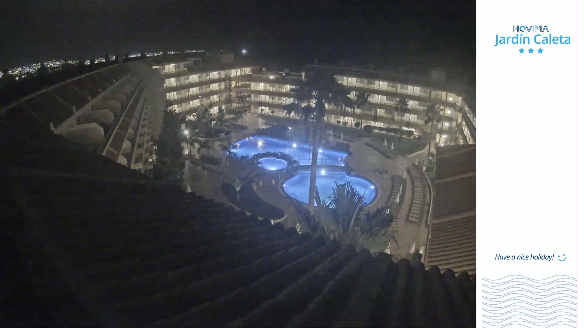 A nighttime aerial view shows a multi-story building complex surrounding a lit swimming pool area, with palm trees visible.