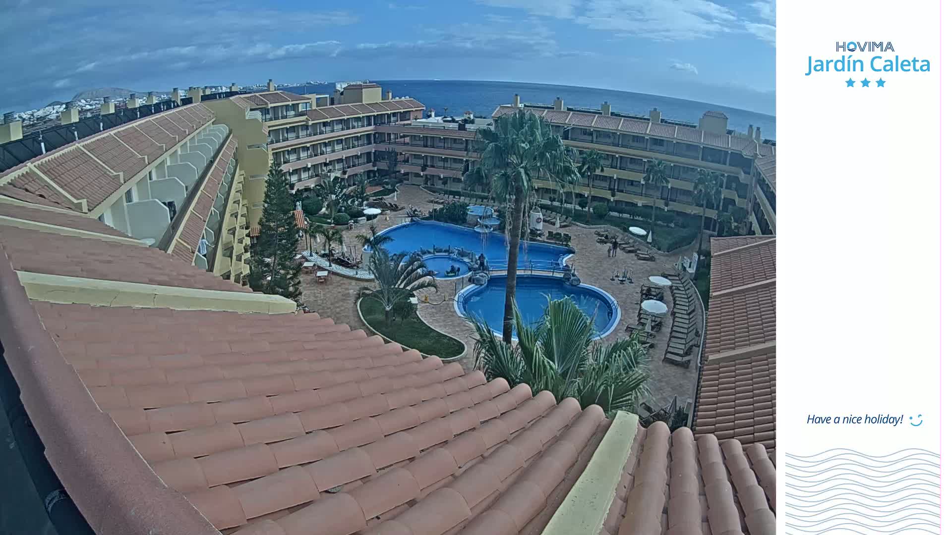 An overhead view of a resort with a large swimming pool, under partly cloudy skies and overlooking the ocean.