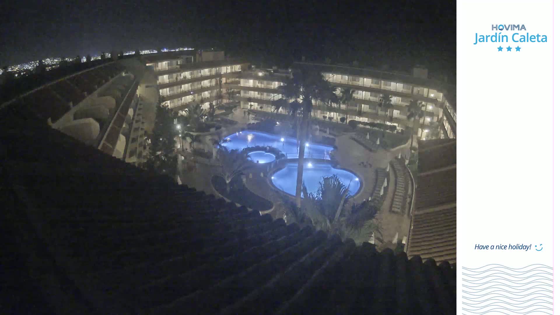 A nighttime aerial view shows a multi-story building complex surrounding a lit swimming pool area with palm trees.