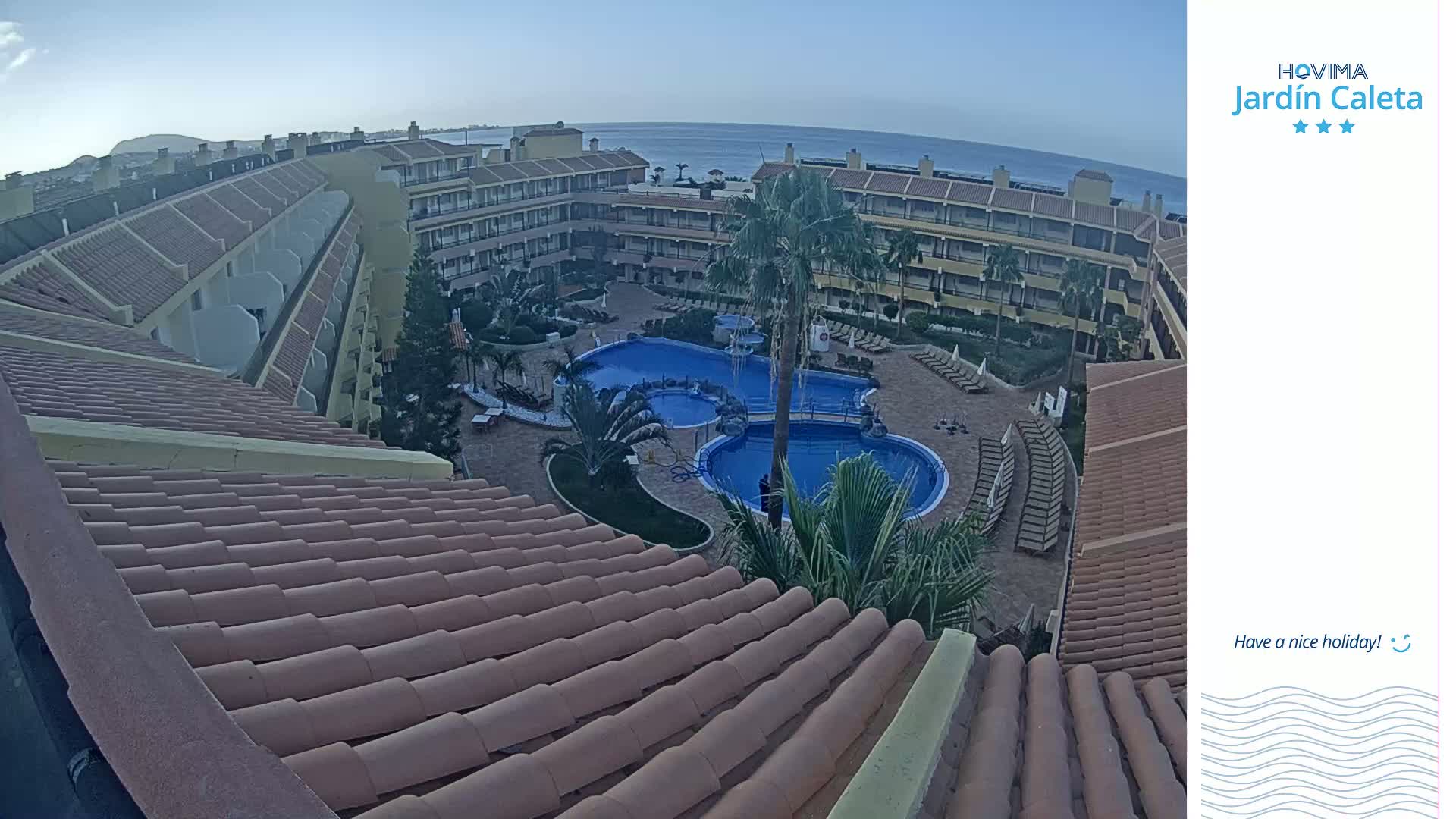 An aerial view shows a resort with multiple buildings surrounding a large pool under a clear sky.