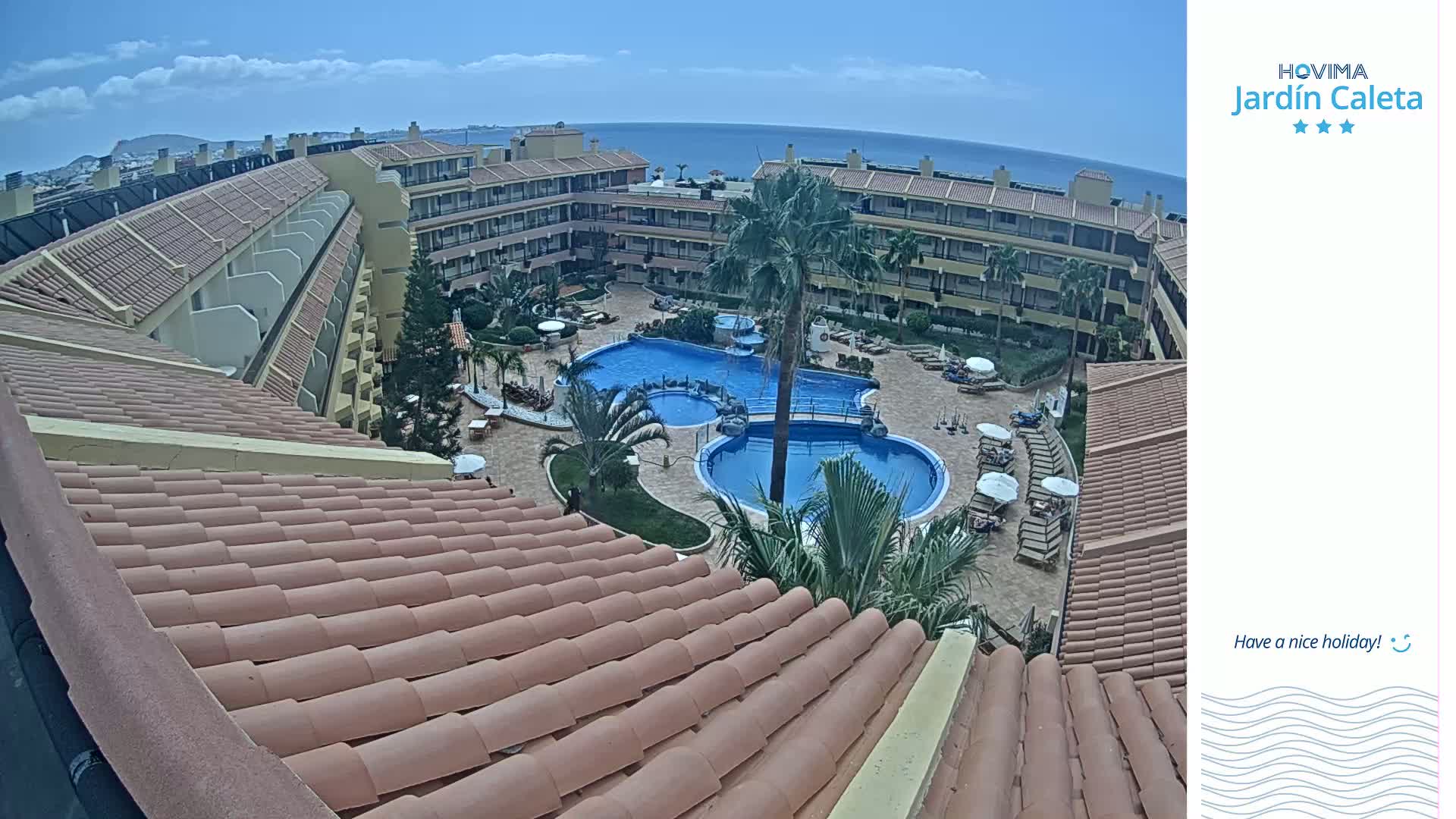 An aerial view shows a resort with a tiled roof, multiple pools, and palm trees under a mostly sunny sky.