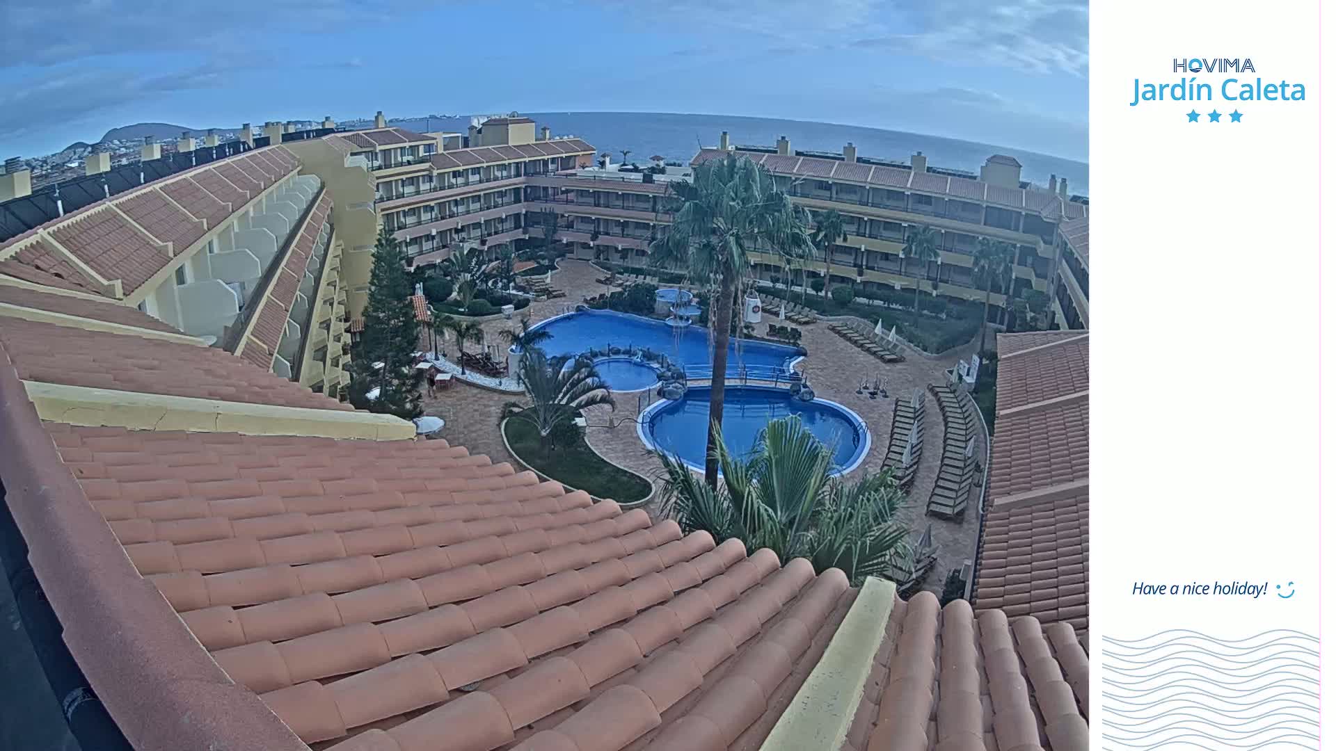 An aerial view shows a resort with a pool and surrounding buildings under a partly cloudy sky.