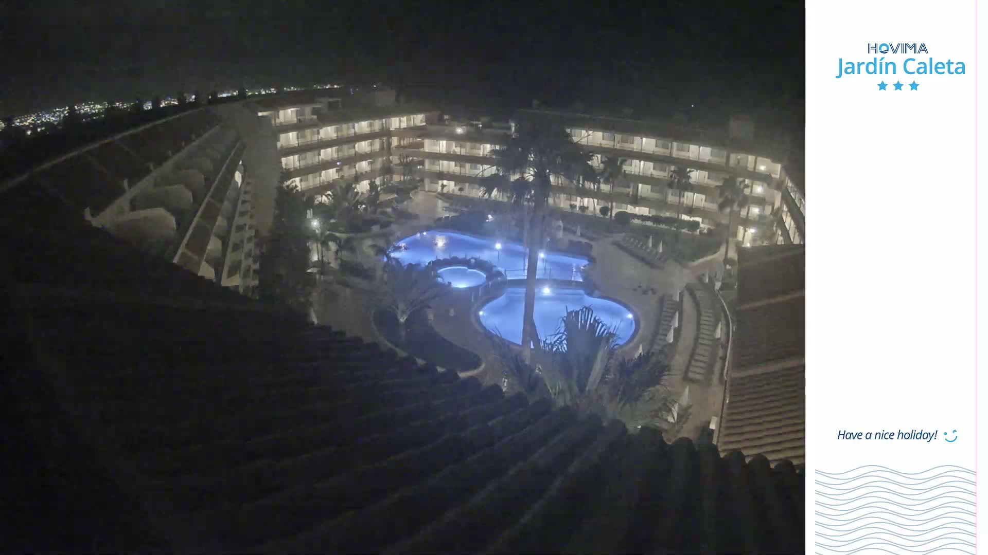 A nighttime, high-angle view shows a multi-story building complex surrounding a lit swimming pool.