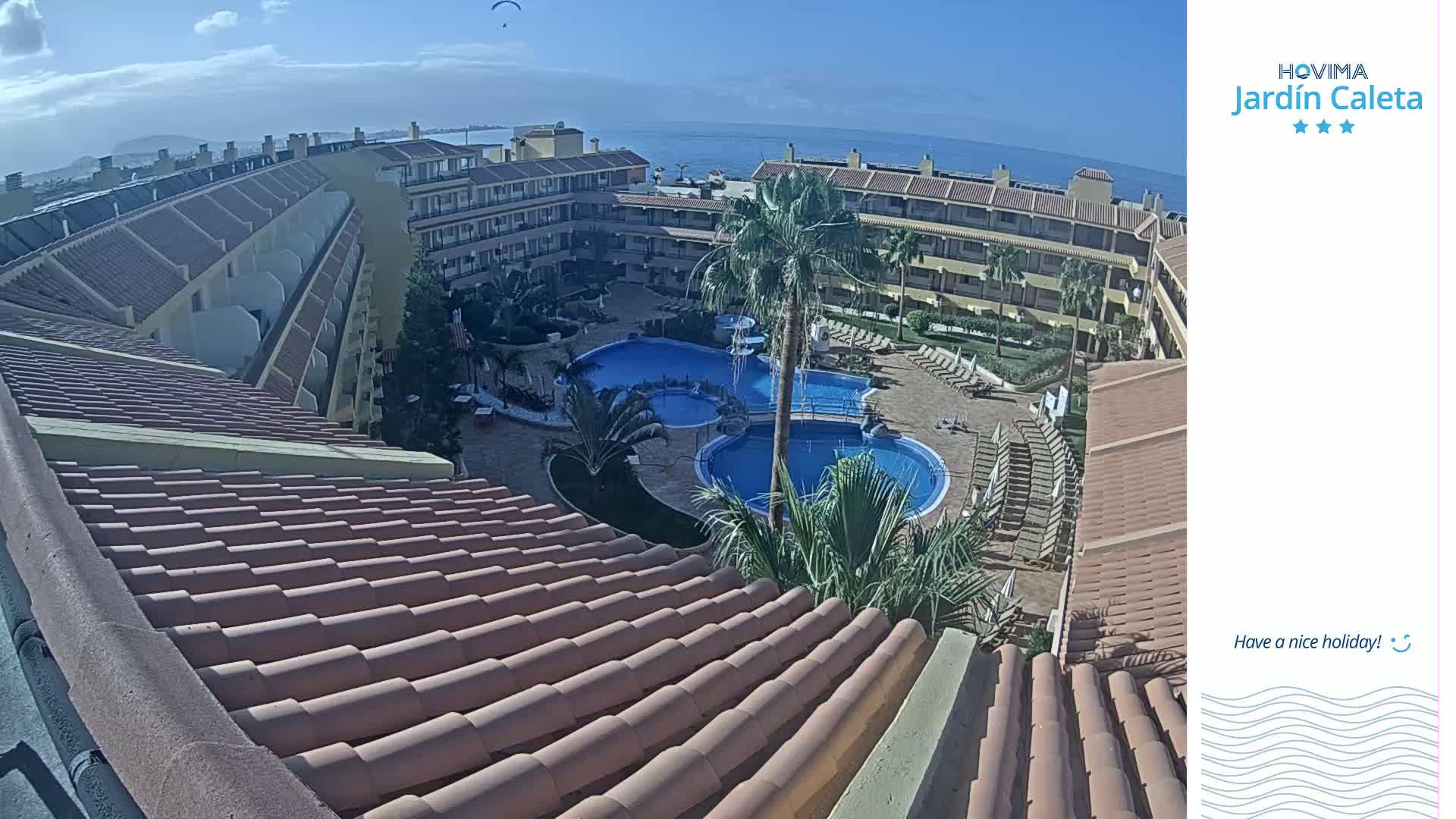 A high-angle view of a resort complex on a sunny day, showcasing multiple buildings surrounding a central pool area with lounge chairs and palm trees, overlooking a body of water in the distance.