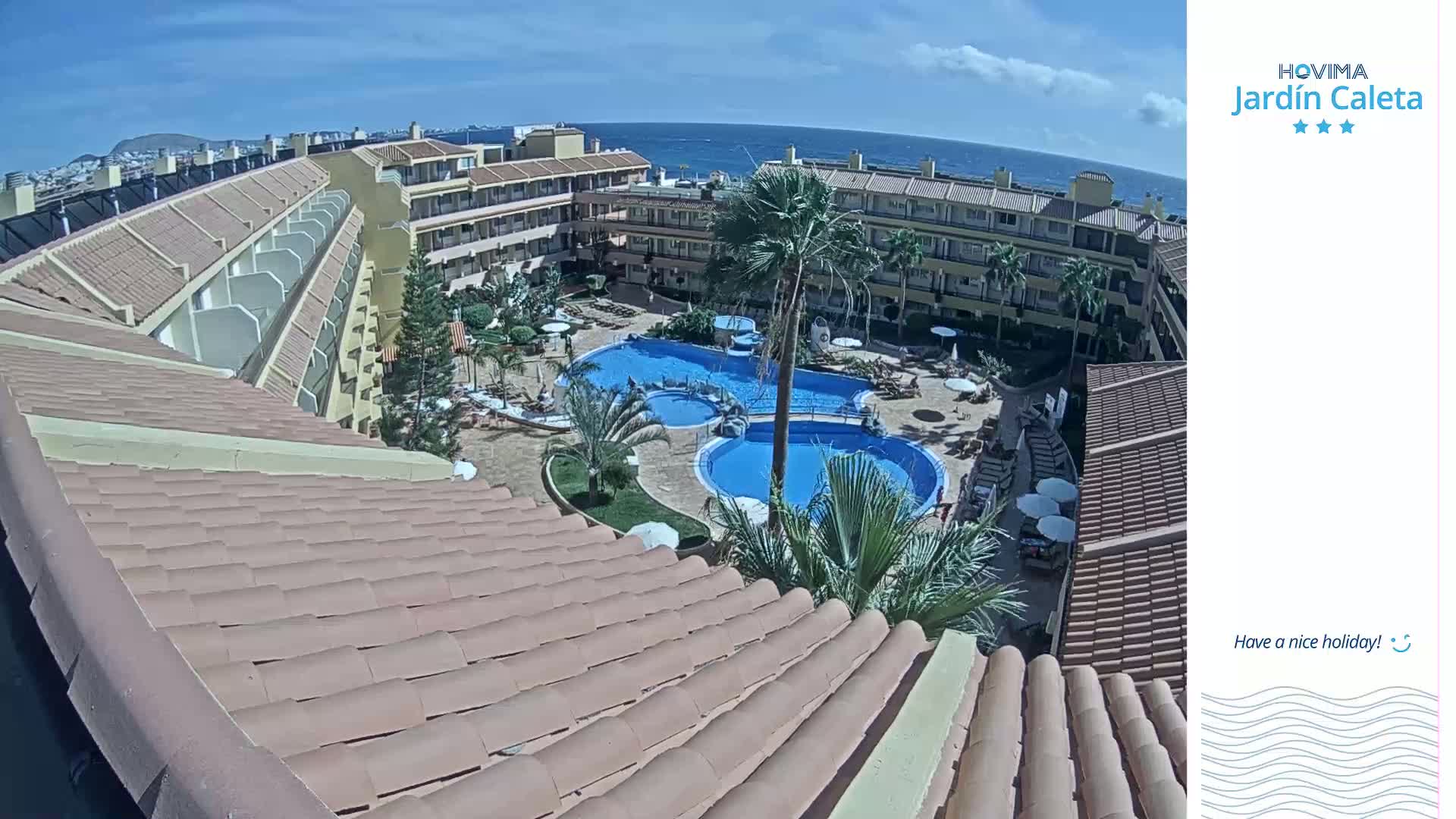 A high-angle view shows a resort with multiple buildings surrounding a multi-tiered swimming pool under a sunny sky.