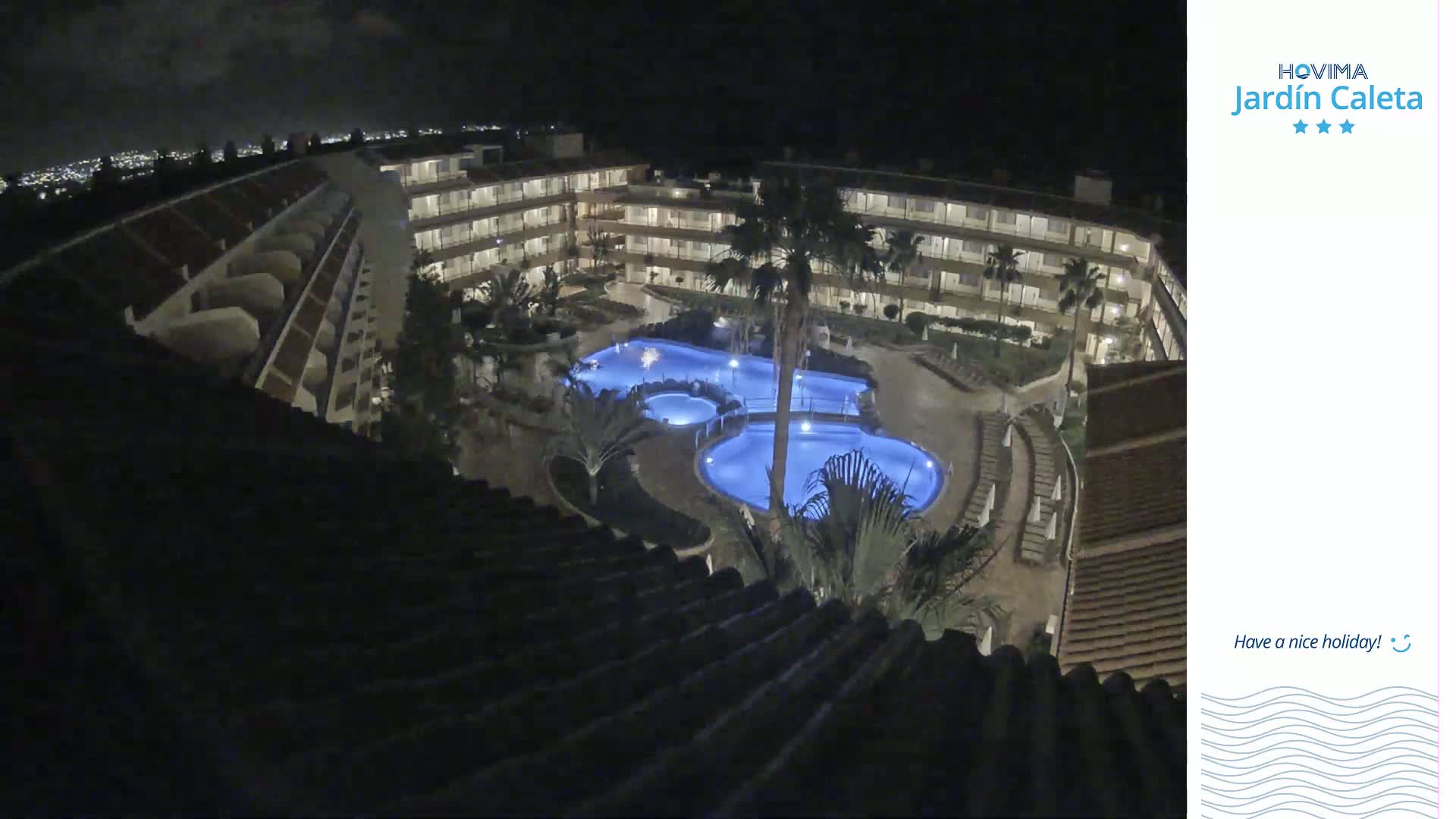A nighttime overhead view shows a multi-story building complex surrounding a lit swimming pool area with palm trees.