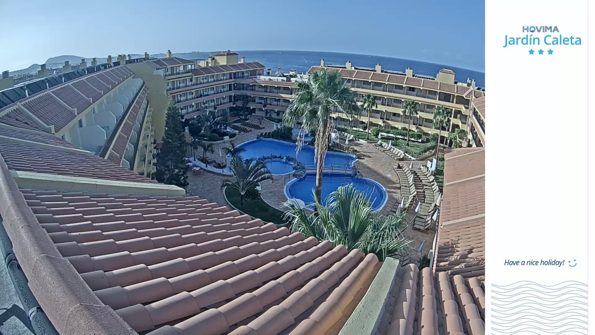 An aerial view of a resort complex with multiple pools surrounded by buildings and palm trees, under sunny skies and overlooking a calm ocean.