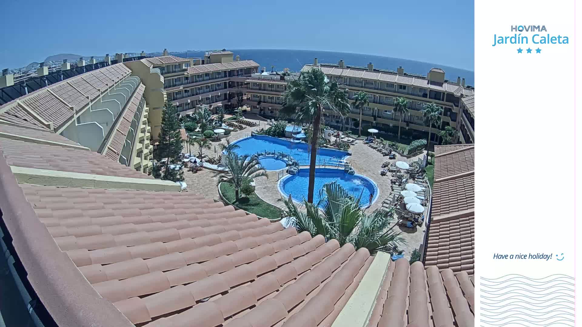 A rooftop view of a resort complex on a sunny day, showcasing a large pool area surrounded by buildings and palm trees, overlooking the ocean in the distance.
