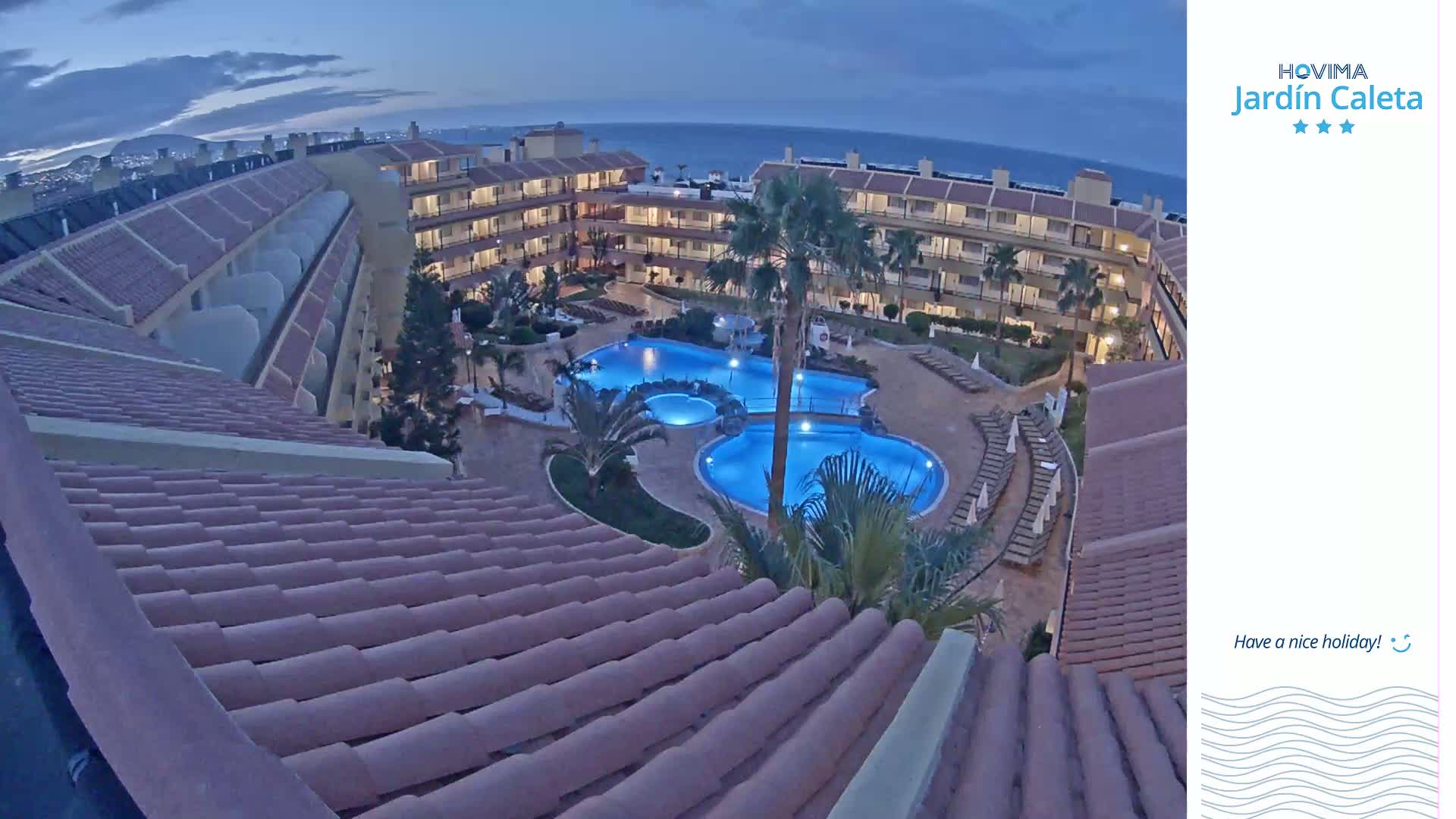 An illuminated resort pool and surrounding buildings are viewed from an adjacent rooftop at dusk.