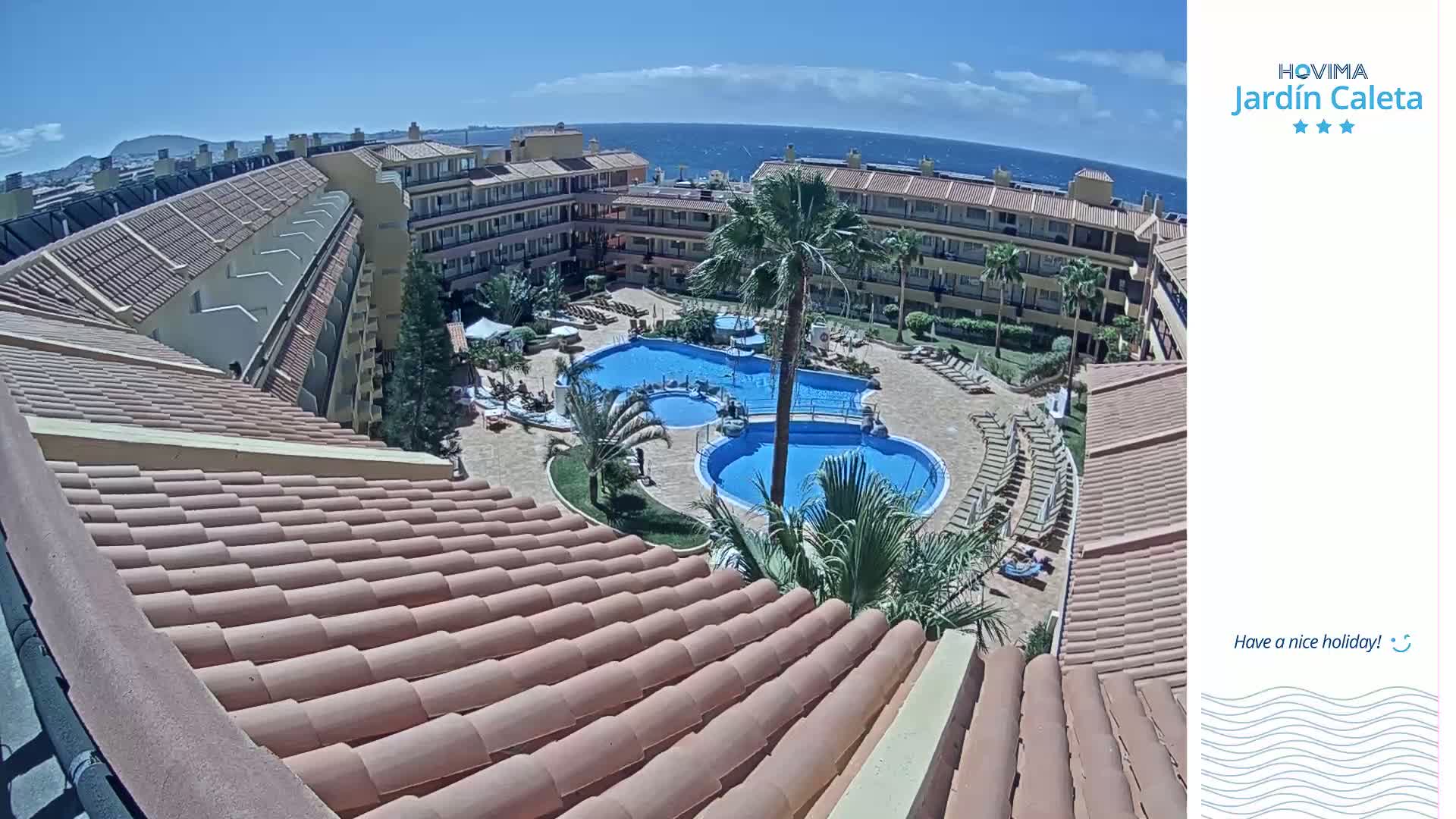 An overhead view shows a resort courtyard with two swimming pools surrounded by lounge chairs and palm trees on a sunny day, next to the ocean.
