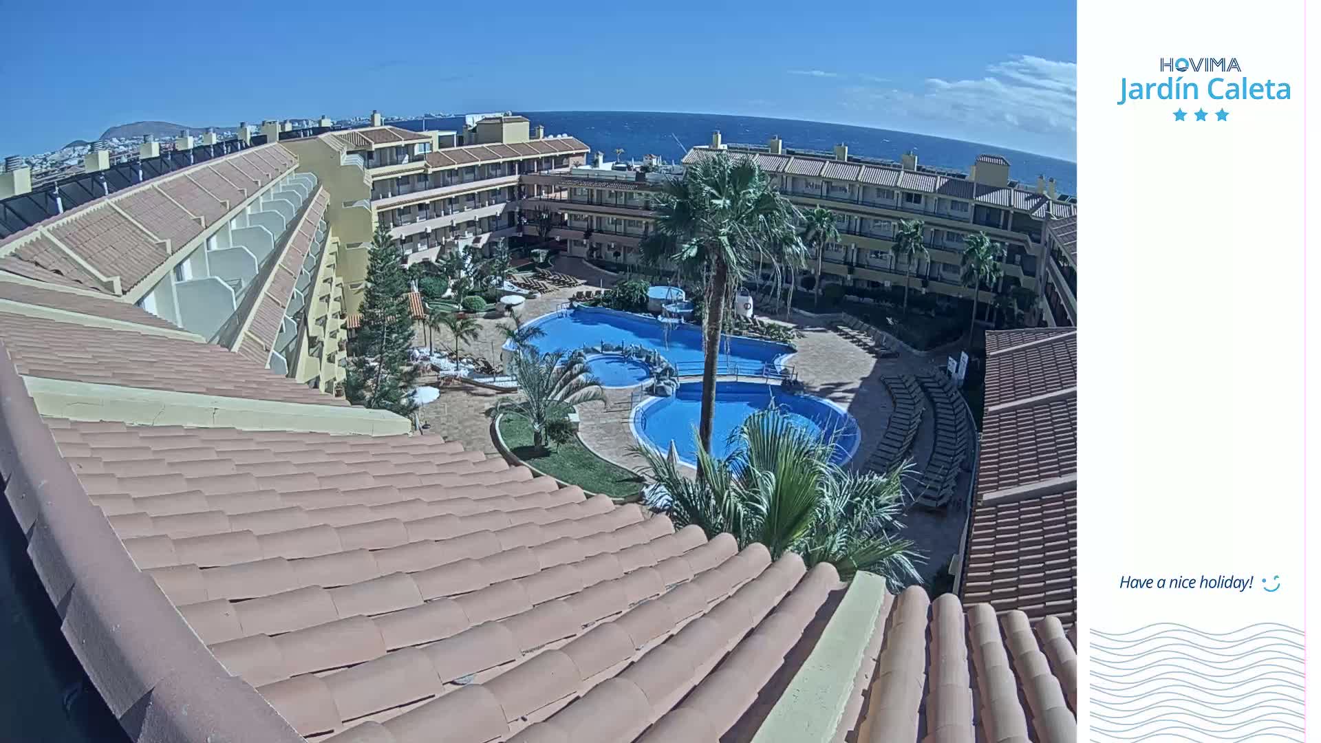 An aerial view shows a resort complex with multiple buildings, swimming pools, and palm trees under a clear, sunny sky overlooking a calm ocean.
