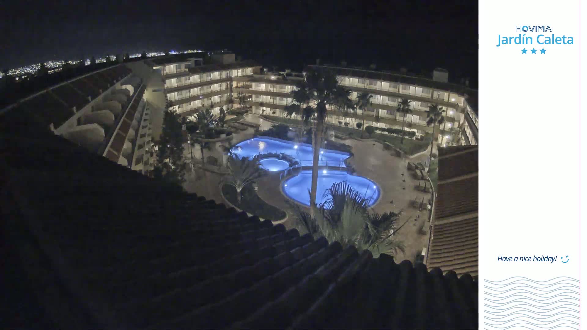 A nighttime aerial view shows a multi-story building complex surrounding a lit, kidney-shaped swimming pool.