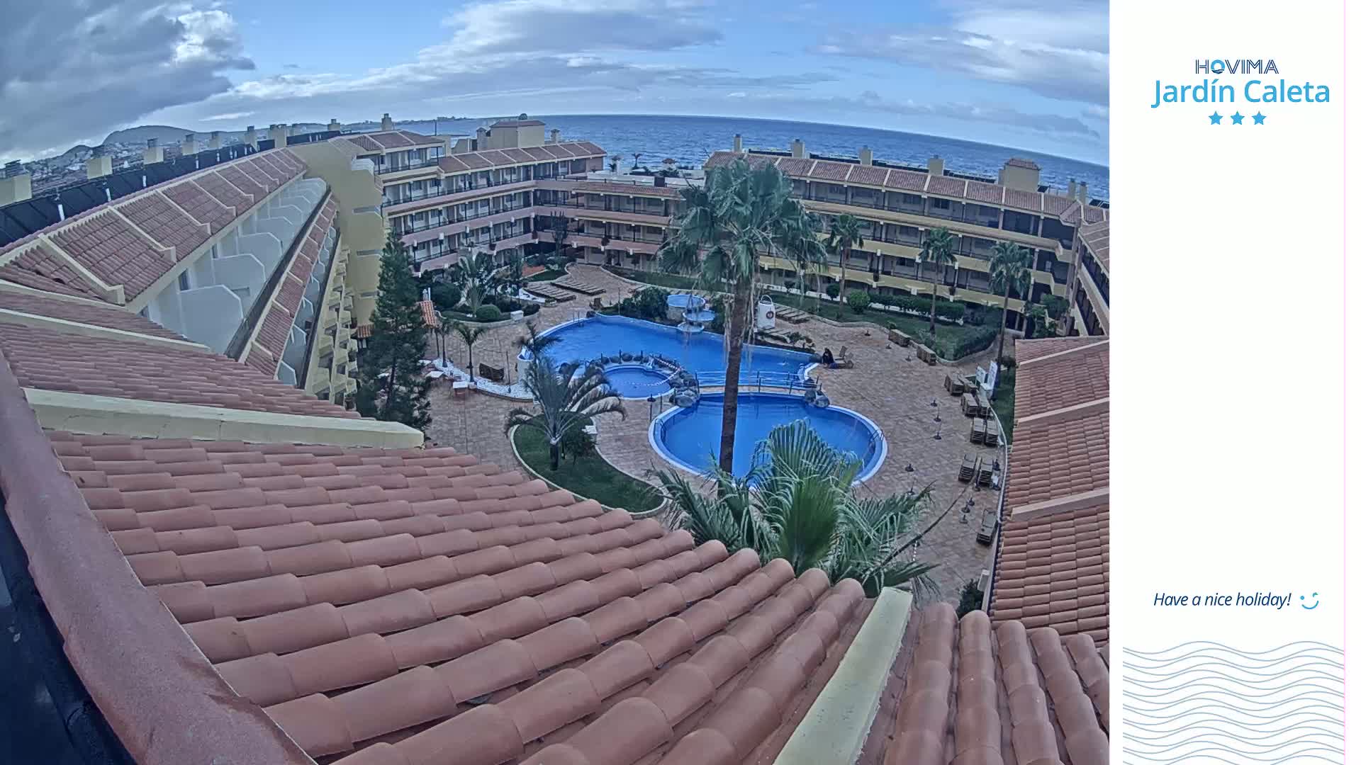 An overhead view shows a courtyard with two swimming pools surrounded by a multi-story building under a partly cloudy sky.