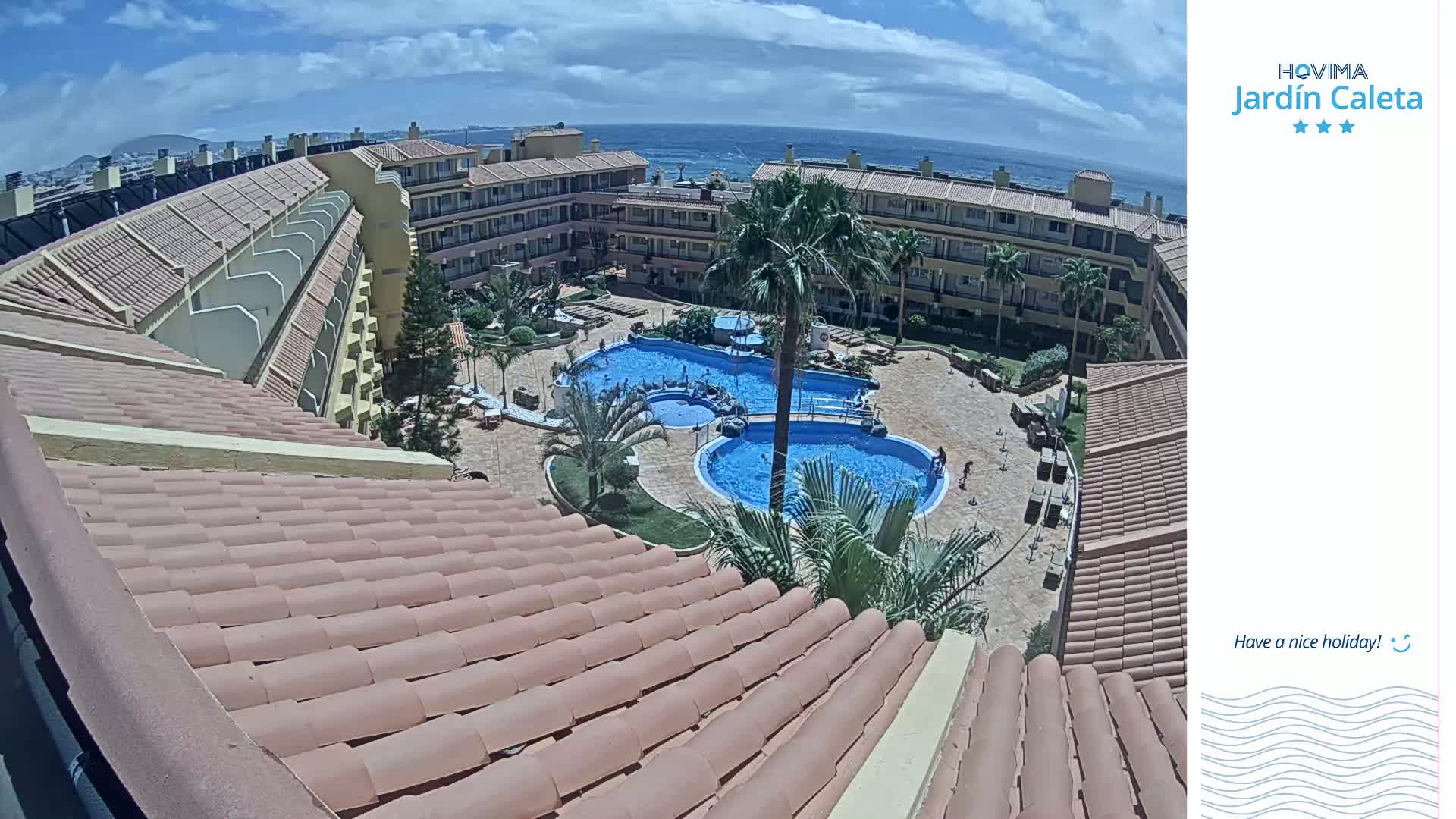 A tiled roof overlooks a sunny courtyard with two swimming pools and a low-rise building complex bordering the ocean.