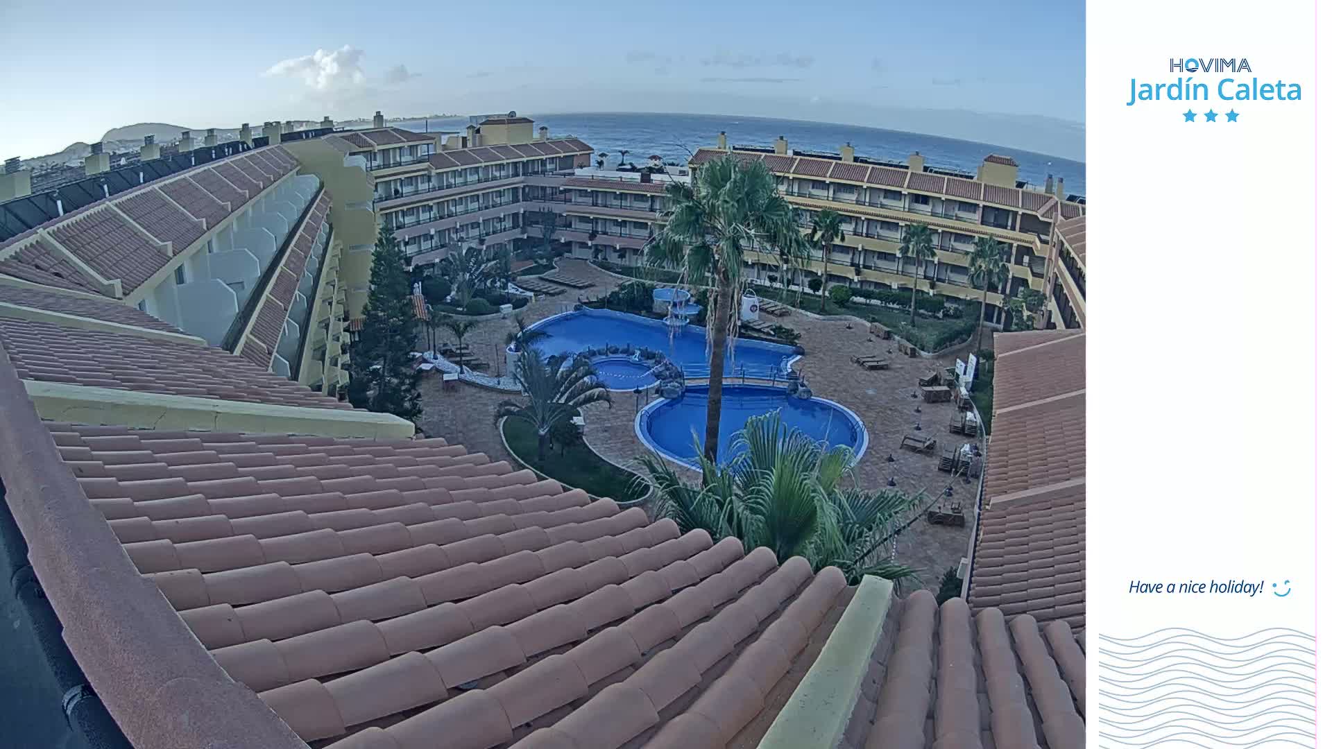 An aerial view of a resort complex with a pool and surrounding buildings under a mostly clear sky.