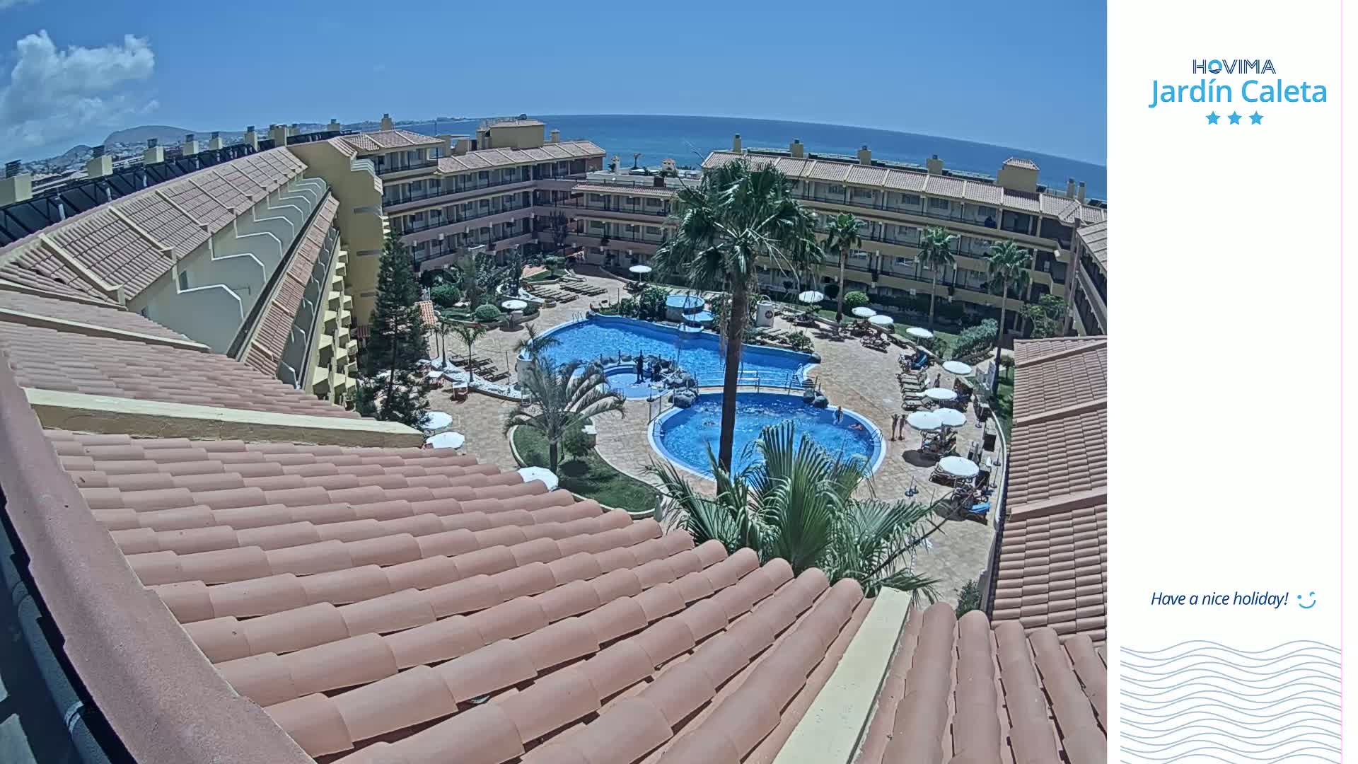 A sunny day overlooks a resort's courtyard with two swimming pools surrounded by buildings and palm trees.