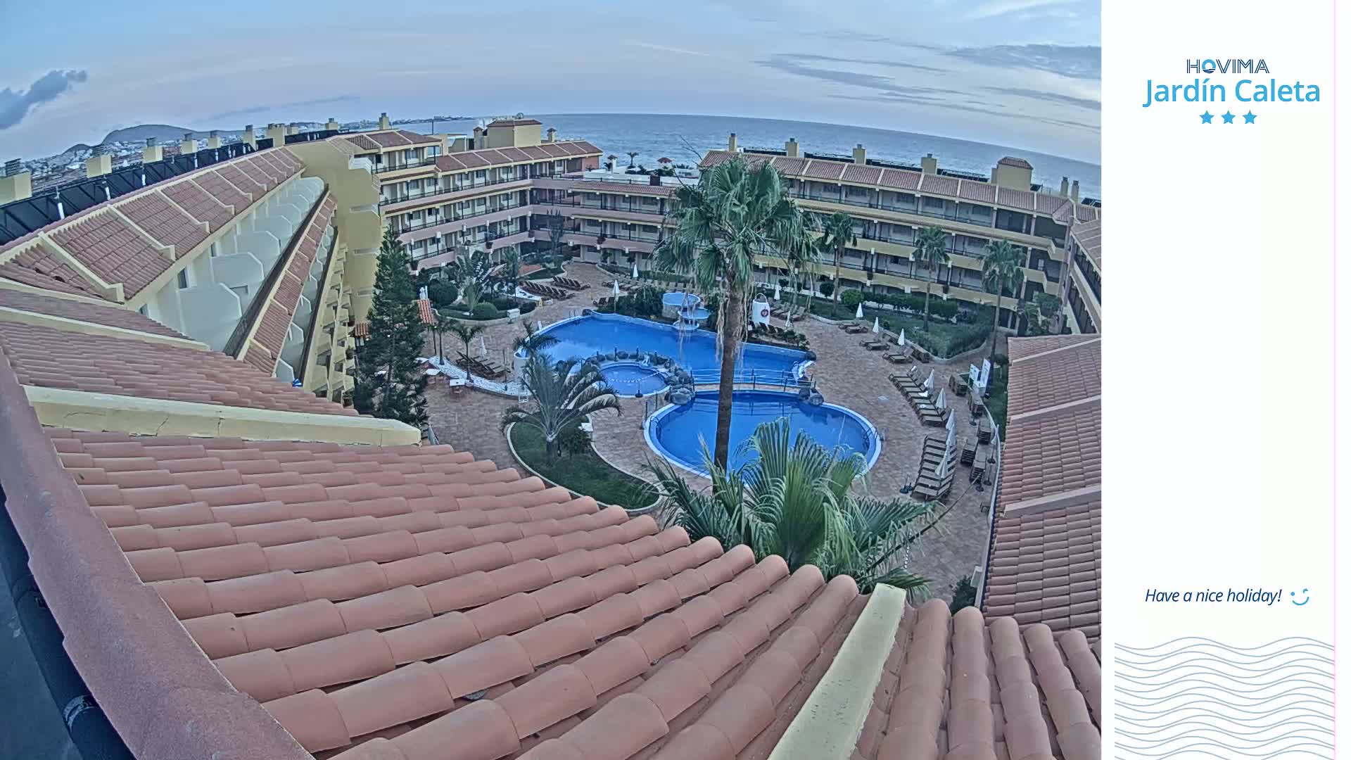 An aerial view of a resort complex with a tiled roof, two swimming pools, and palm trees, overlooking the ocean under a partly cloudy sky.
