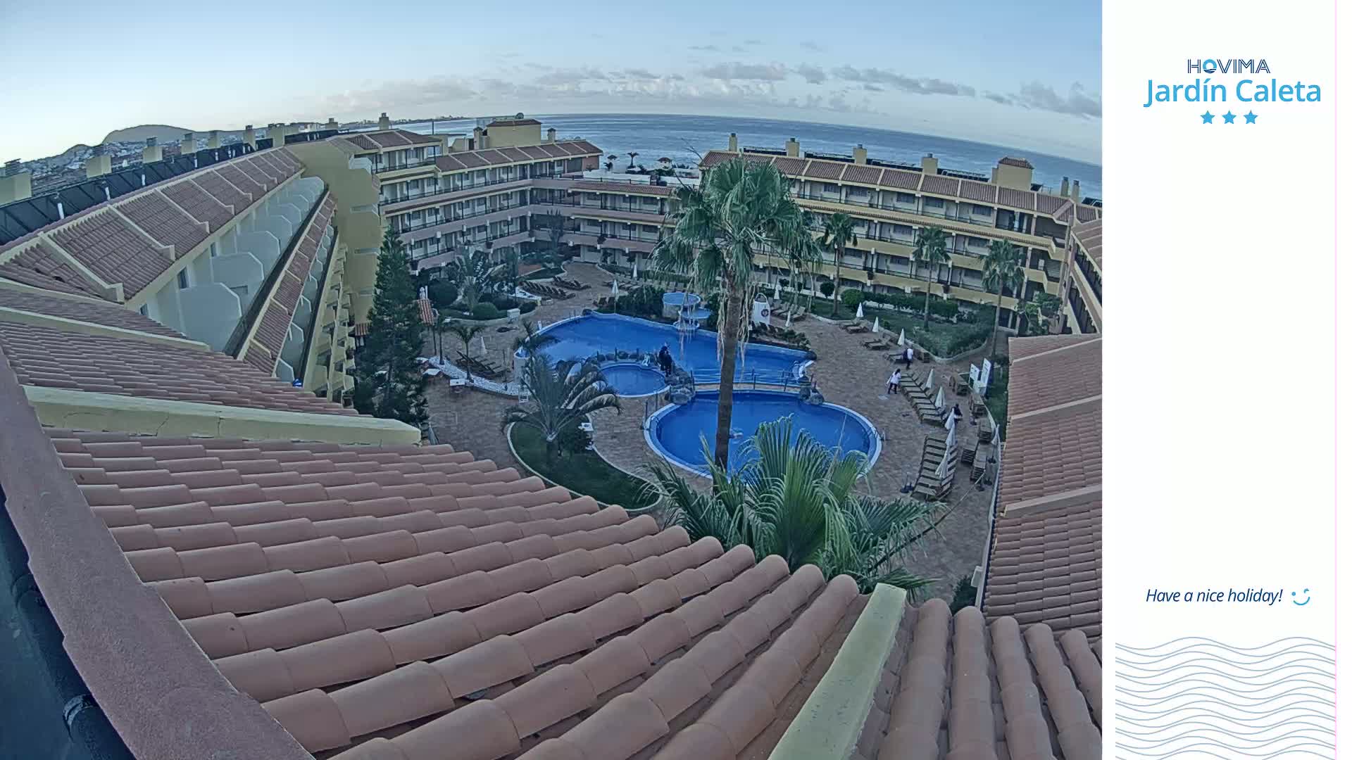 An aerial view shows a resort with a multi-level building surrounding a courtyard with two swimming pools under a clear, twilight sky.