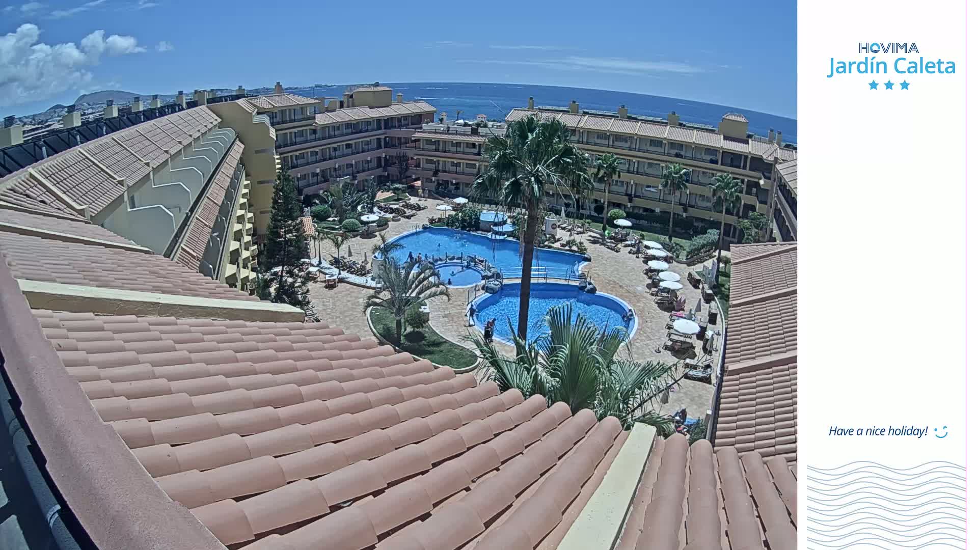 An overhead view of a resort complex with multiple swimming pools and surrounding buildings, under sunny skies and overlooking a calm ocean.
