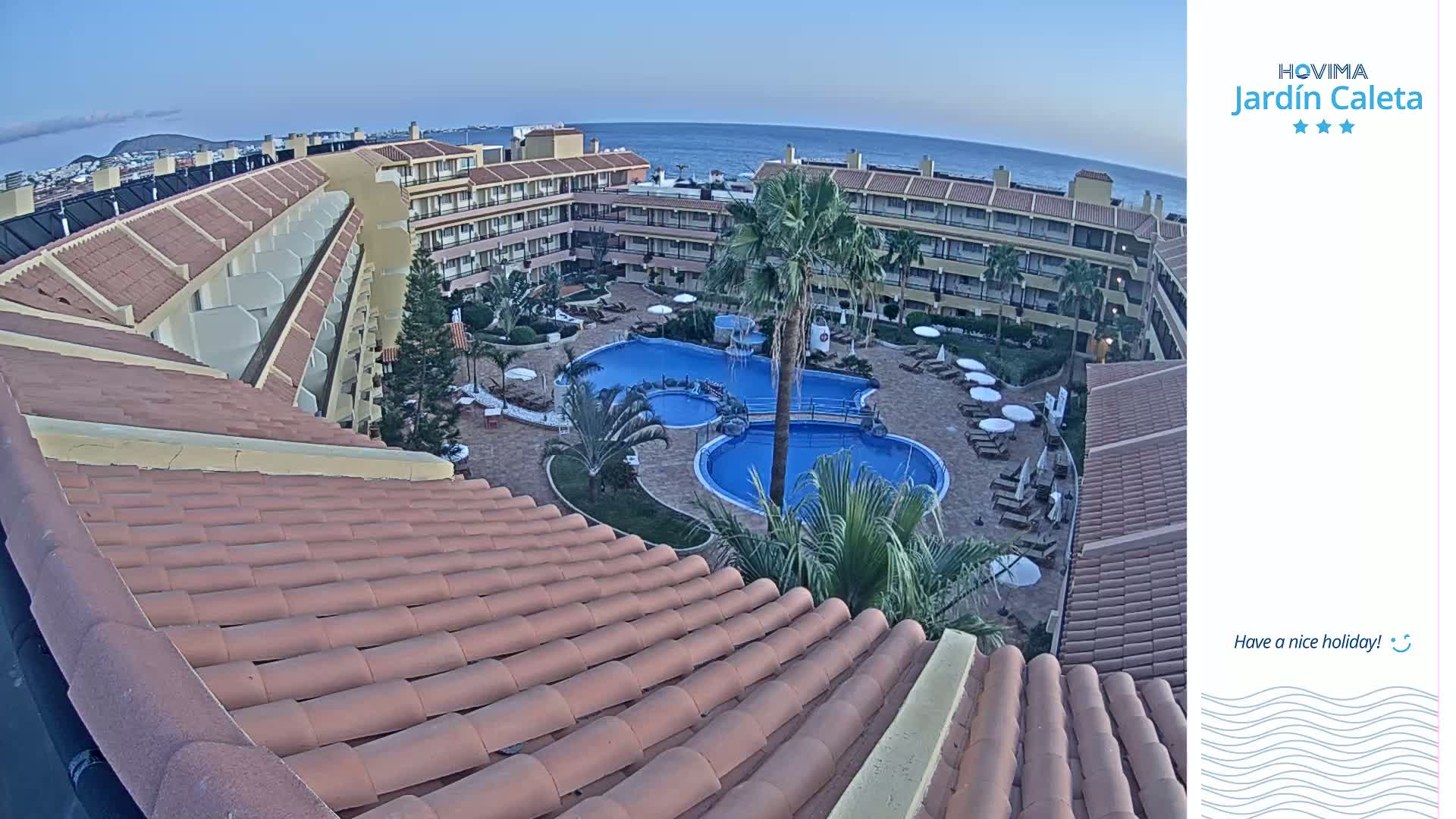 An aerial view shows a resort with multiple buildings surrounding a swimming pool complex under a clear evening sky.