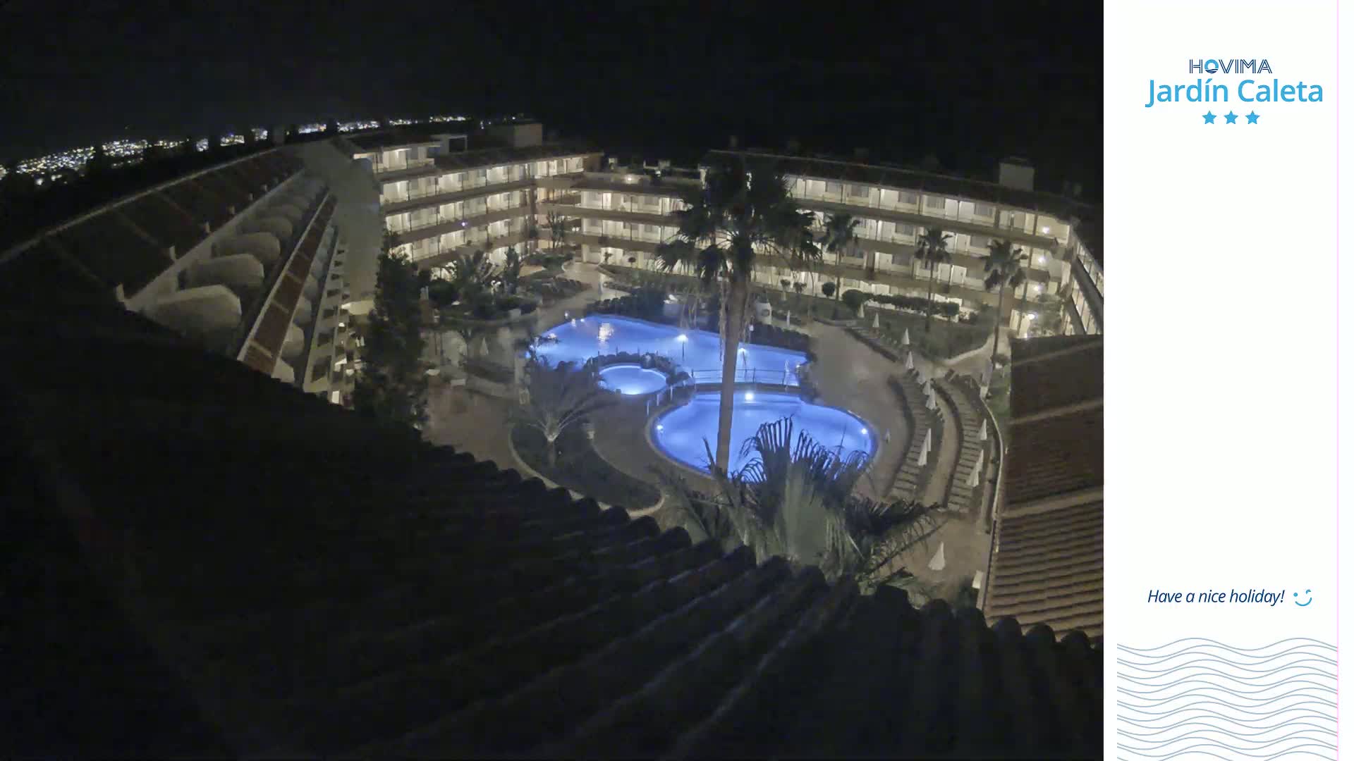 A nighttime aerial view shows a multi-story hotel complex surrounding a lit, multi-level swimming pool.