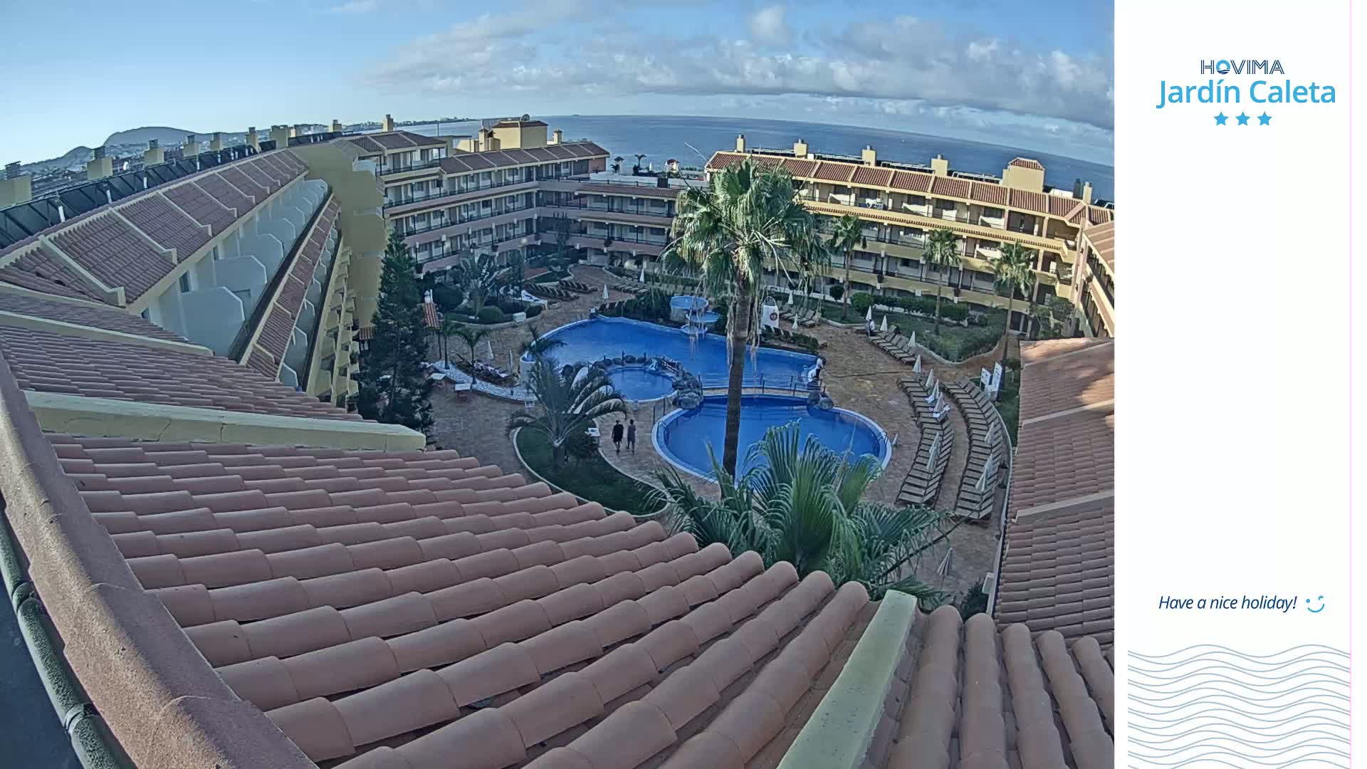 A tiled roof overlooks a courtyard with a swimming pool and a multi-story building under a partly cloudy sky.