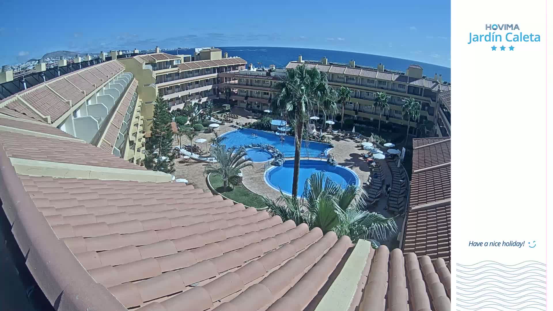 A sunny day overlooking a resort's pool area with multiple buildings, palm trees, and a view of the ocean in the distance.