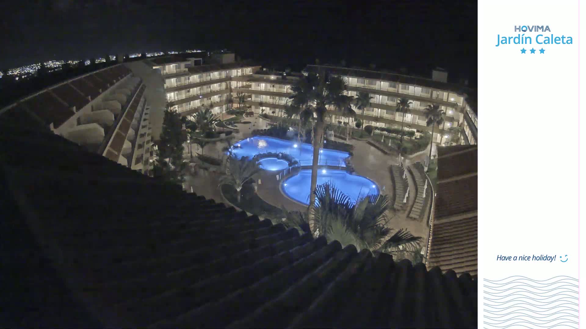 A nighttime, high-angle view shows a multi-story building surrounding a complex of illuminated swimming pools.