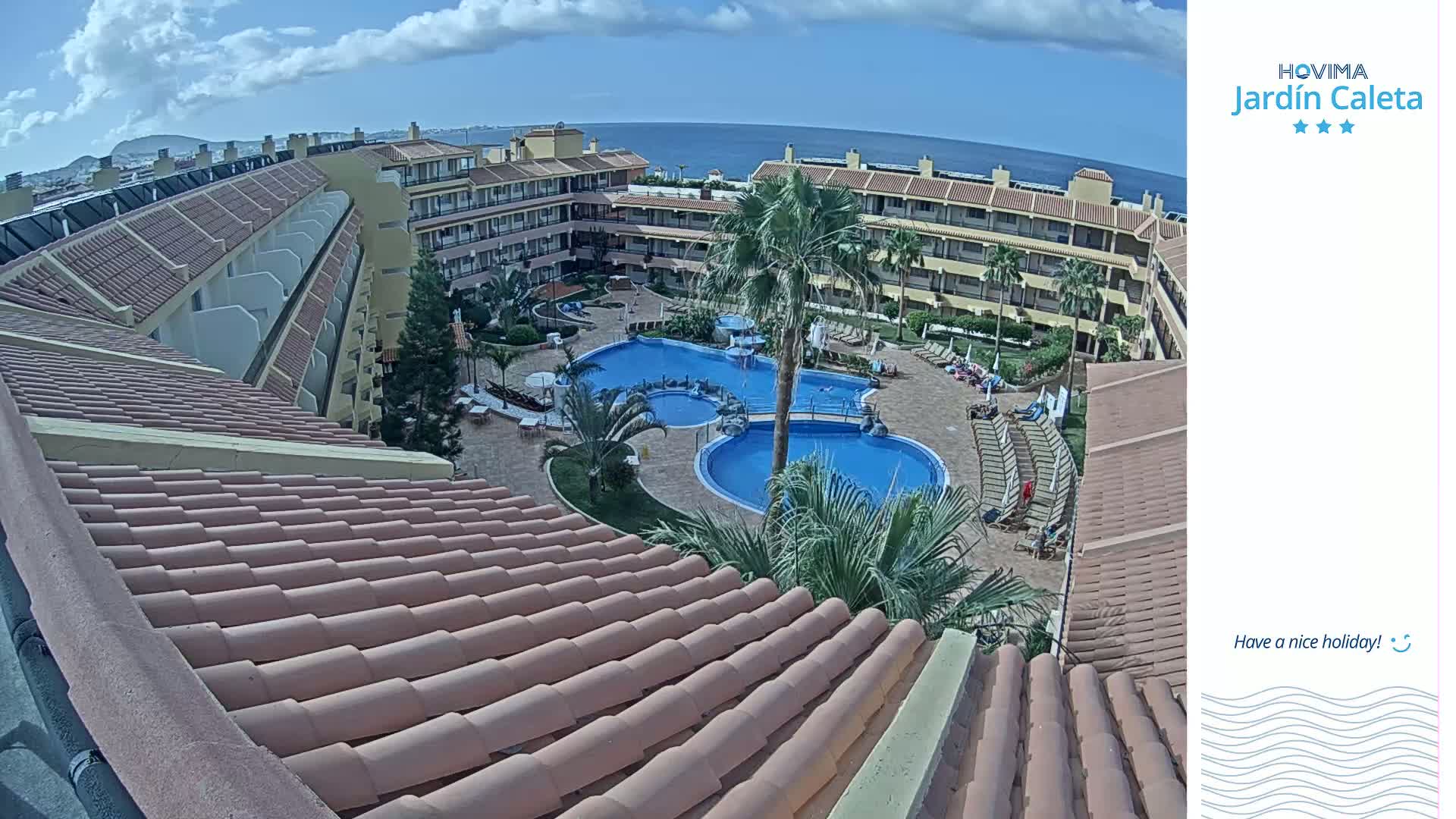 A resort's courtyard with multiple pools, surrounded by buildings under a partly cloudy sky, is viewed from an upper-level roof.