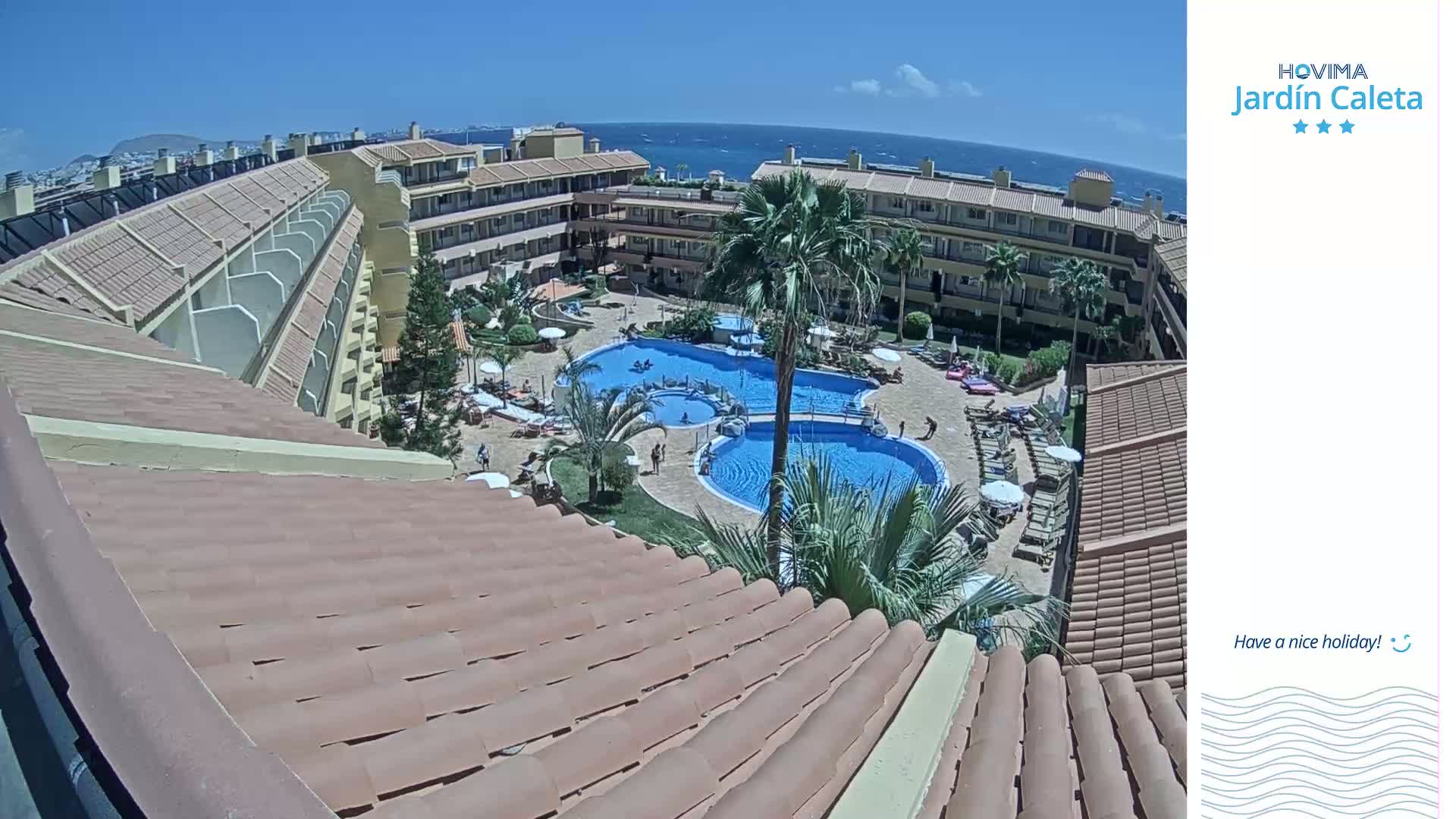 A sunny view overlooking a resort courtyard with two swimming pools surrounded by lounge chairs and buildings, and a distant ocean.