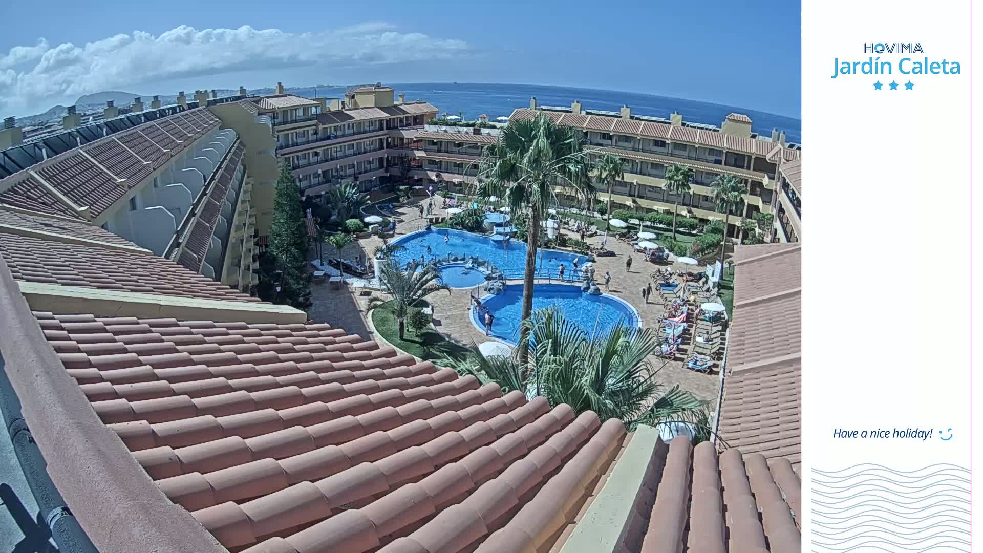 A sunny day overlooking a resort pool area with multiple buildings, palm trees, and ocean views.