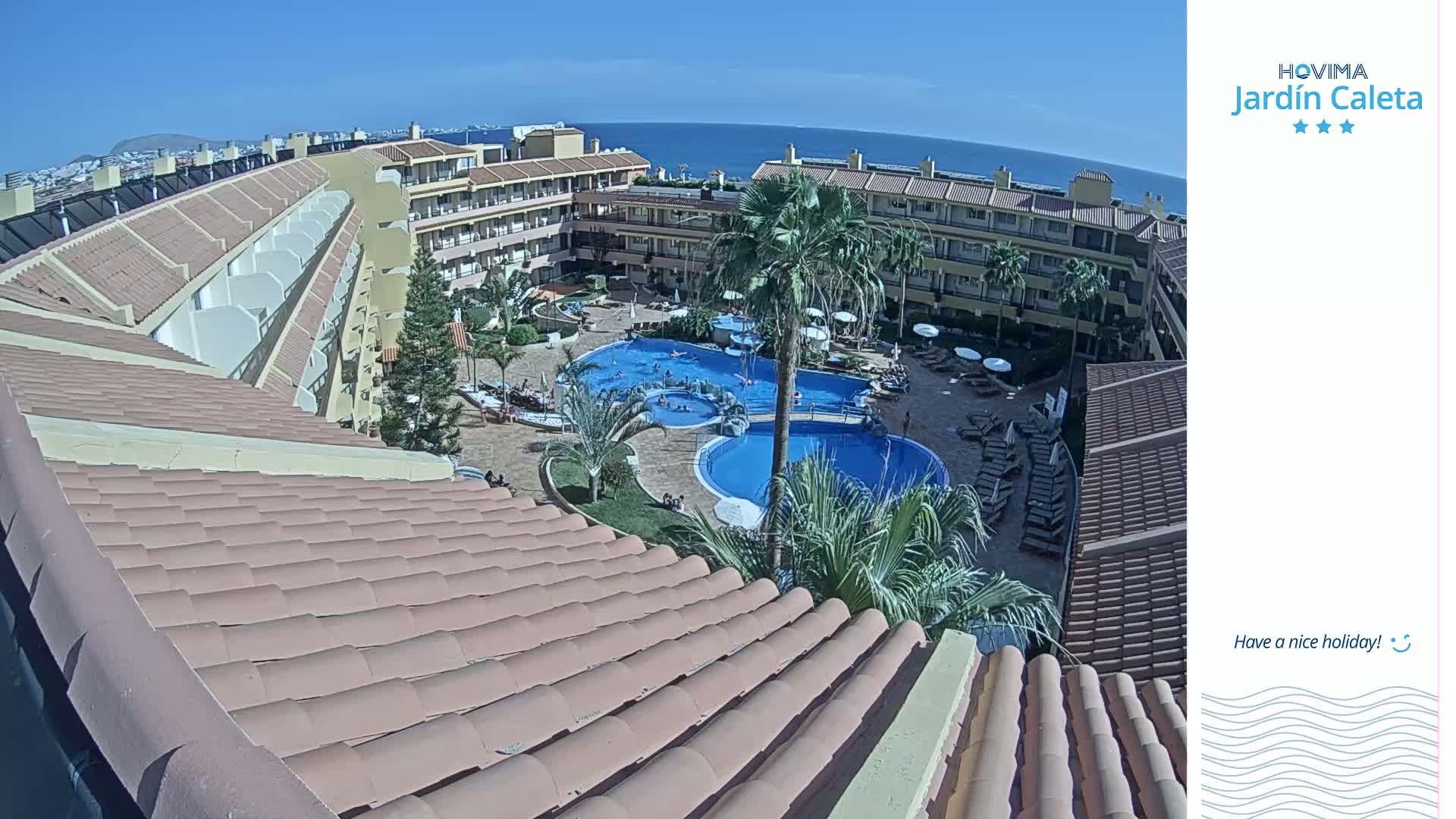 A sunny view from a rooftop shows a resort courtyard with multiple pools surrounded by palm trees and buildings.