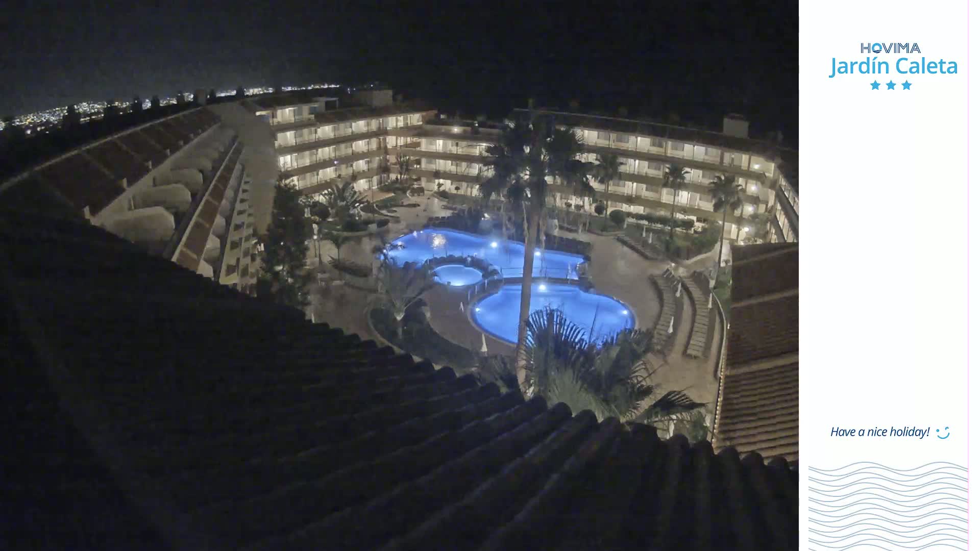 A nighttime, high-angle view shows a multi-story building complex surrounding a lit swimming pool area with palm trees.