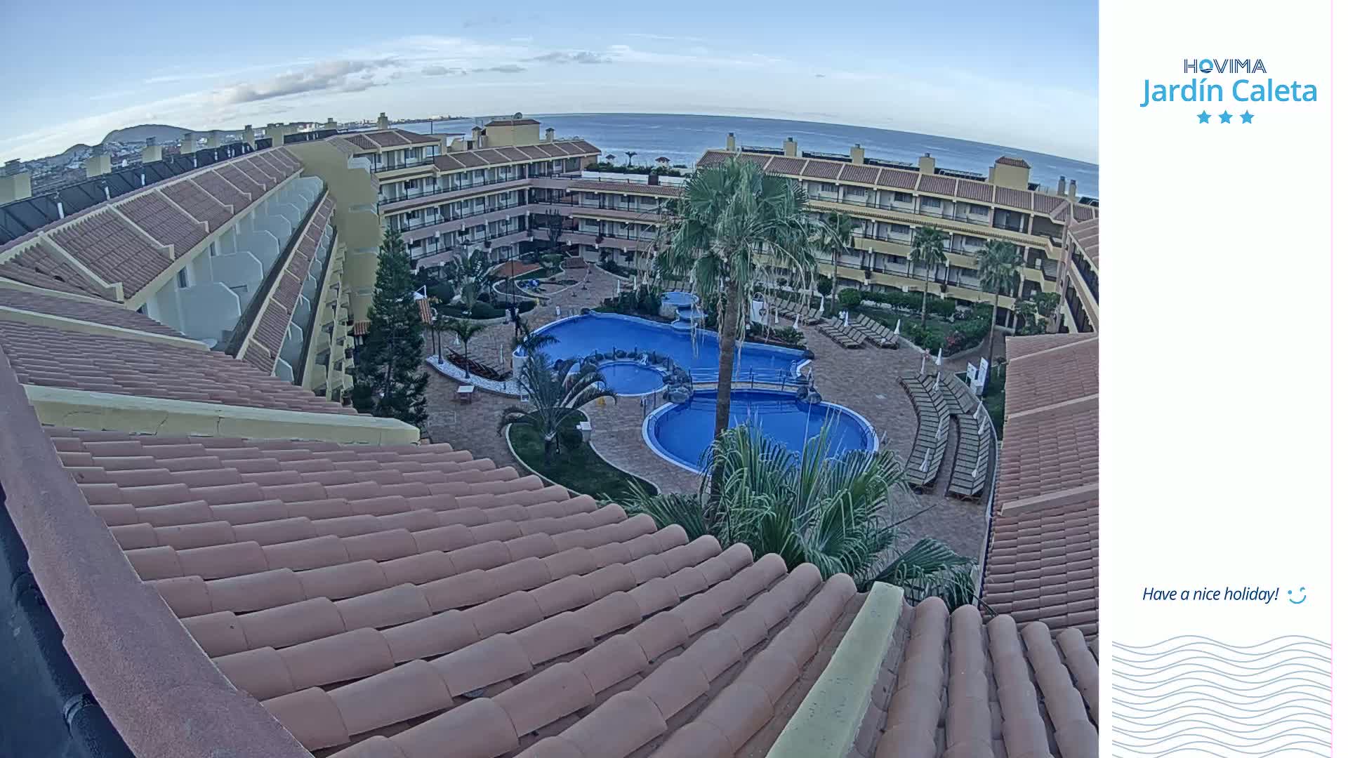 An aerial view shows a resort complex with a swimming pool under a mostly clear sky, overlooking the ocean.