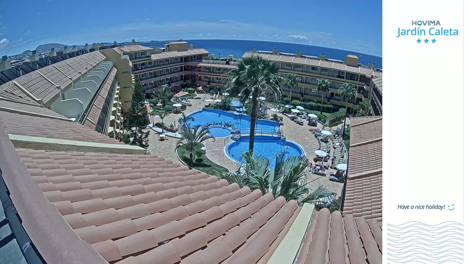 A sunny day overlooking a resort with multiple pools surrounded by buildings and palm trees, next to a calm ocean.