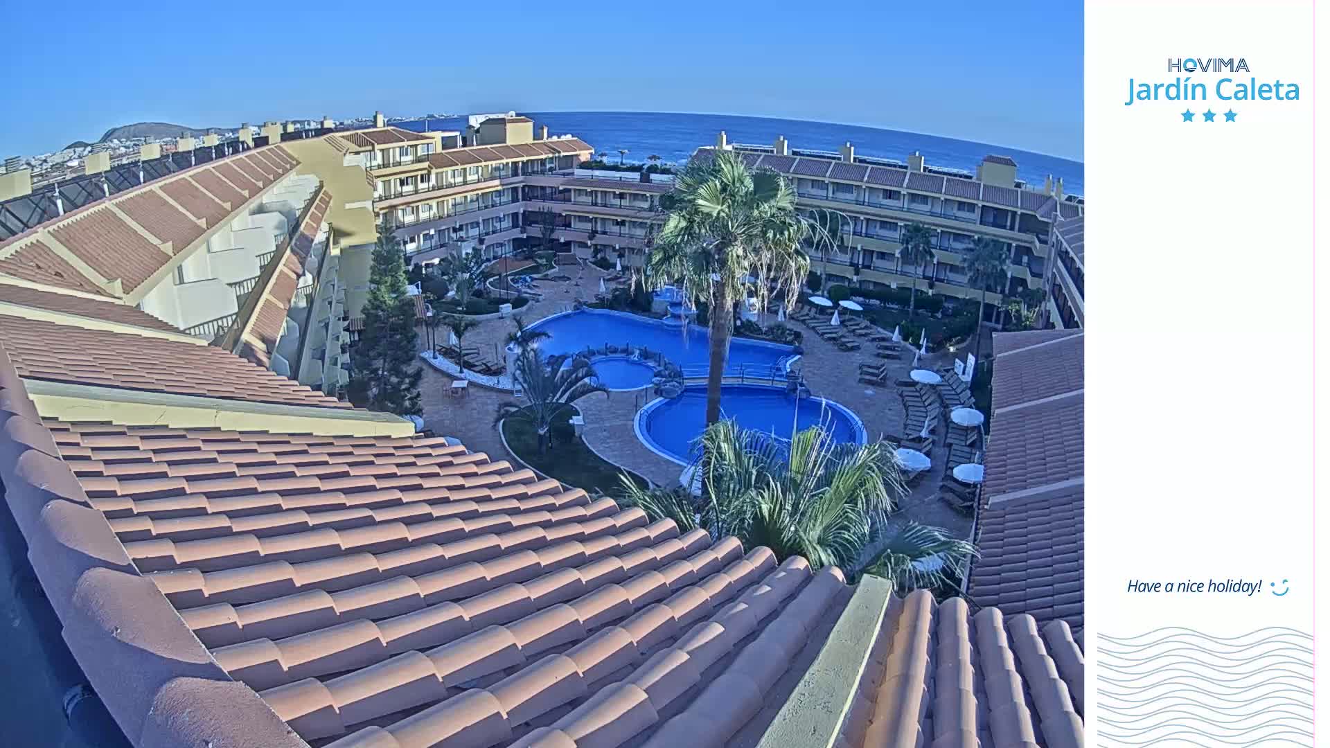 A tiled roof overlooks a resort courtyard with a swimming pool complex, palm trees, and a clear view of the ocean under a sunny sky.