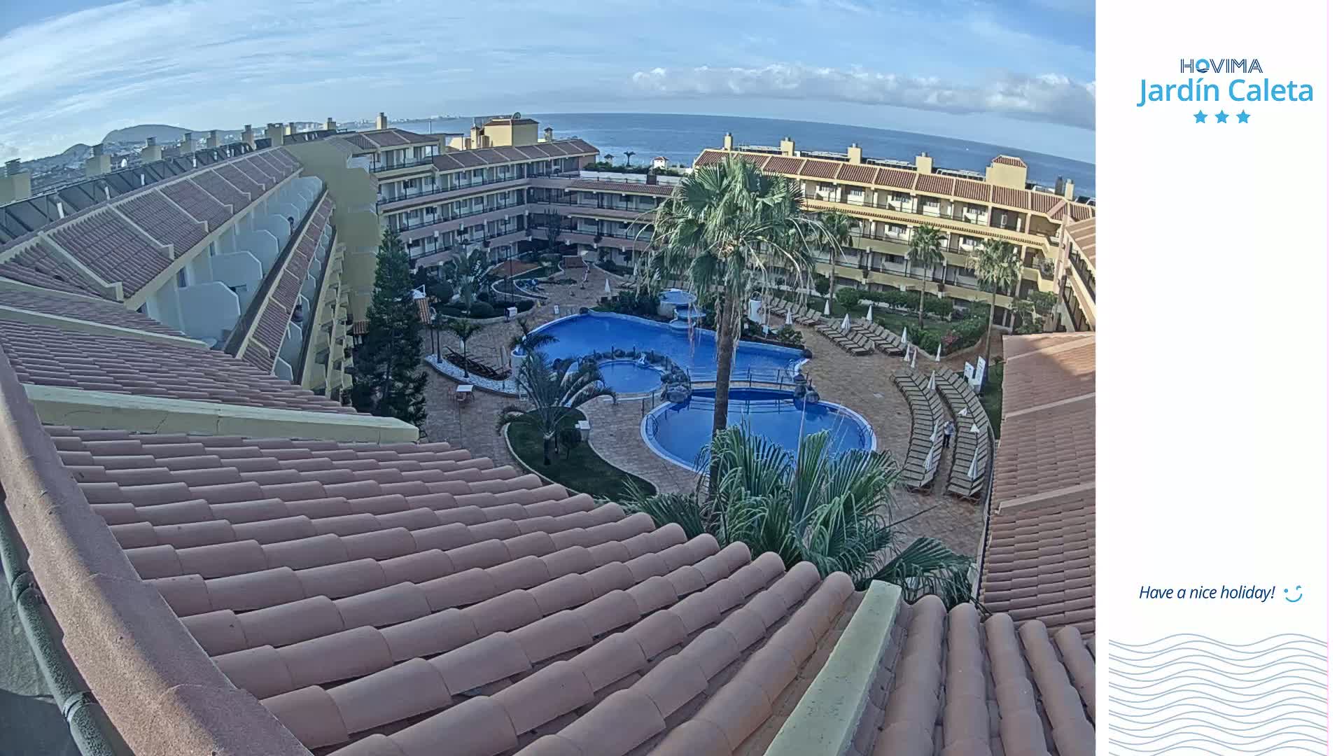 A tiled roof overlooks a courtyard with a swimming pool complex and a multi-story building, all under a mostly clear, sunny sky.