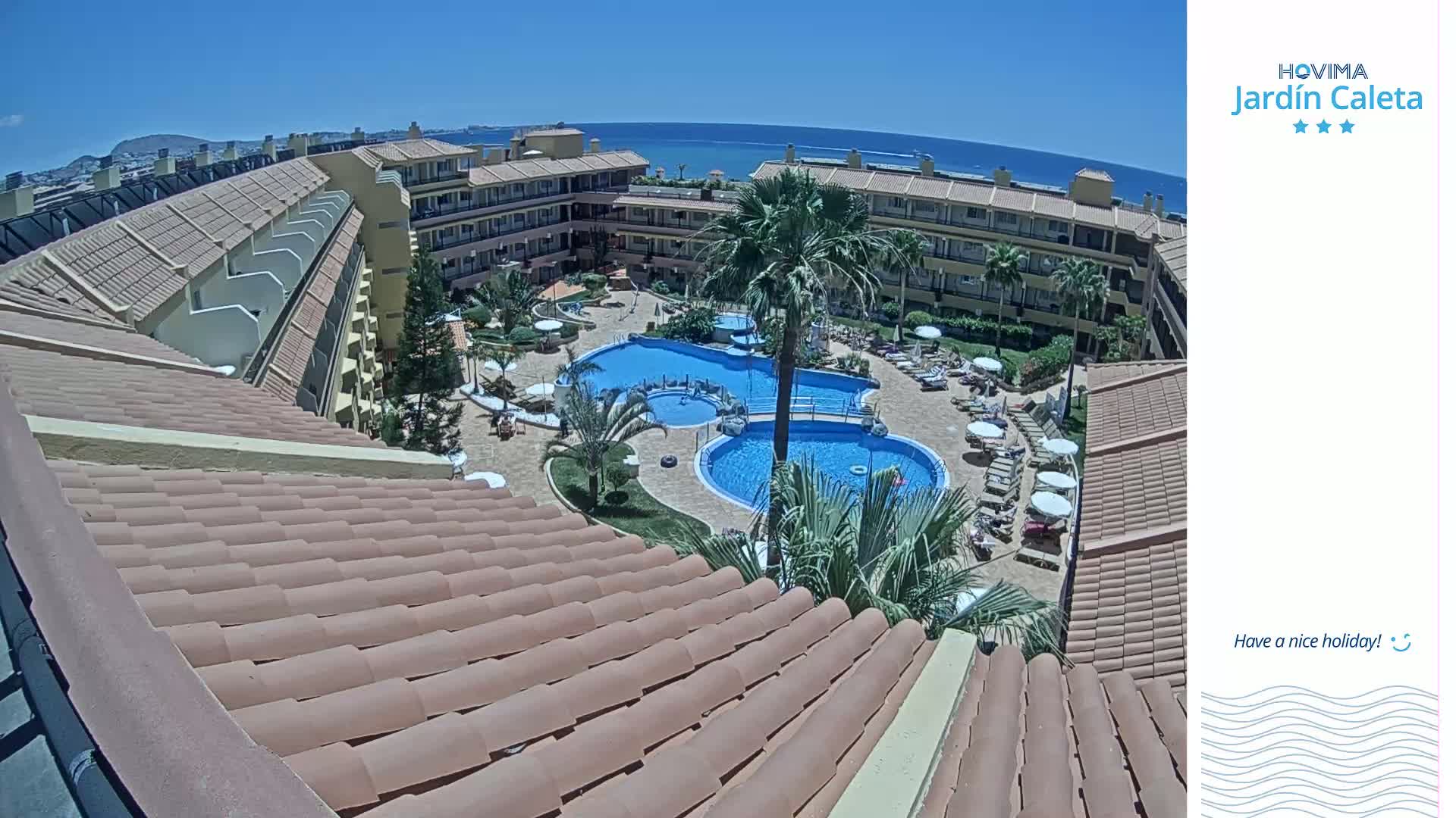 A rooftop view overlooks a resort pool area with multiple pools, lounge chairs, and palm trees on a sunny day, with the ocean visible in the distance.