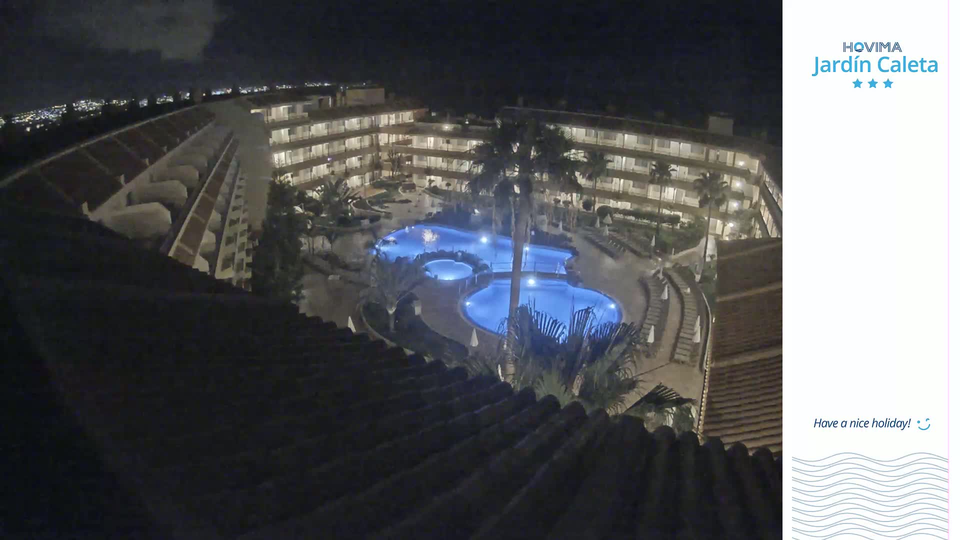 A nighttime, overhead view shows a multi-story hotel complex surrounding a lit, irregularly shaped swimming pool complex.