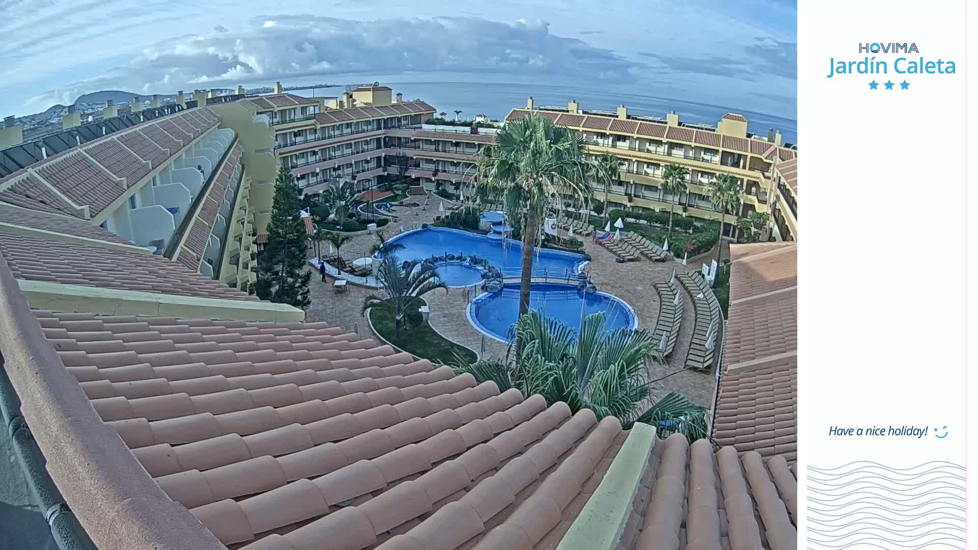 An aerial view shows a multi-story resort building with a large, central swimming pool complex under a partly cloudy sky.