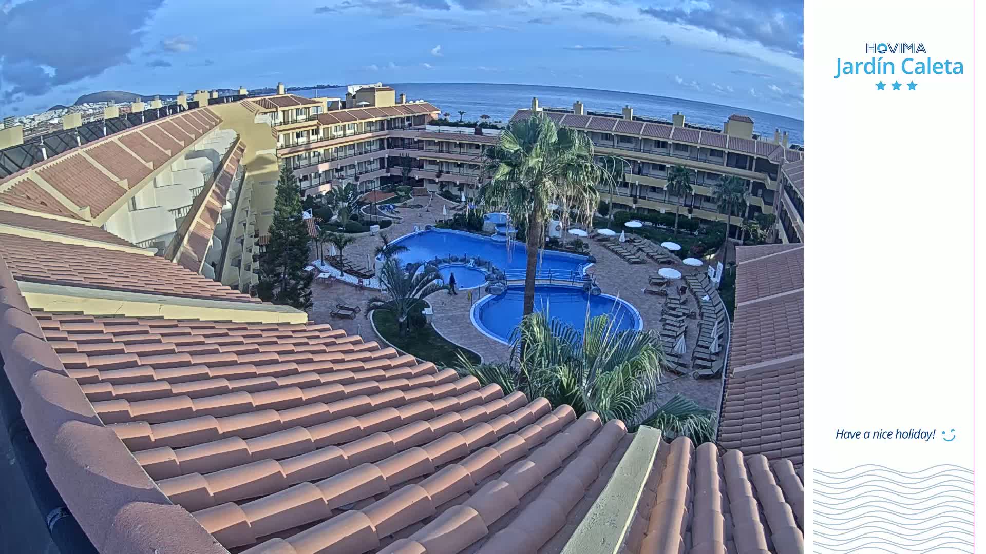 A mostly sunny day shows an aerial view of a resort's pool area, with multiple buildings surrounding a central pool and lounge chairs, all under a partly cloudy sky and overlooking the ocean.