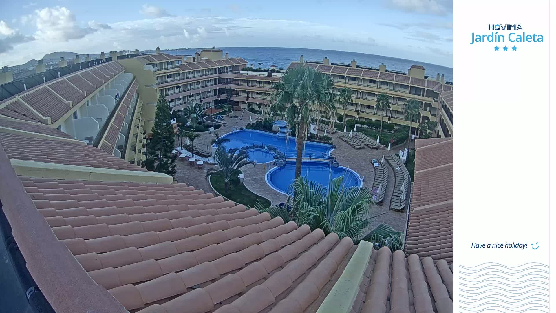 An overhead view shows a resort pool and surrounding buildings under a partly cloudy sky, with the ocean visible in the distance.